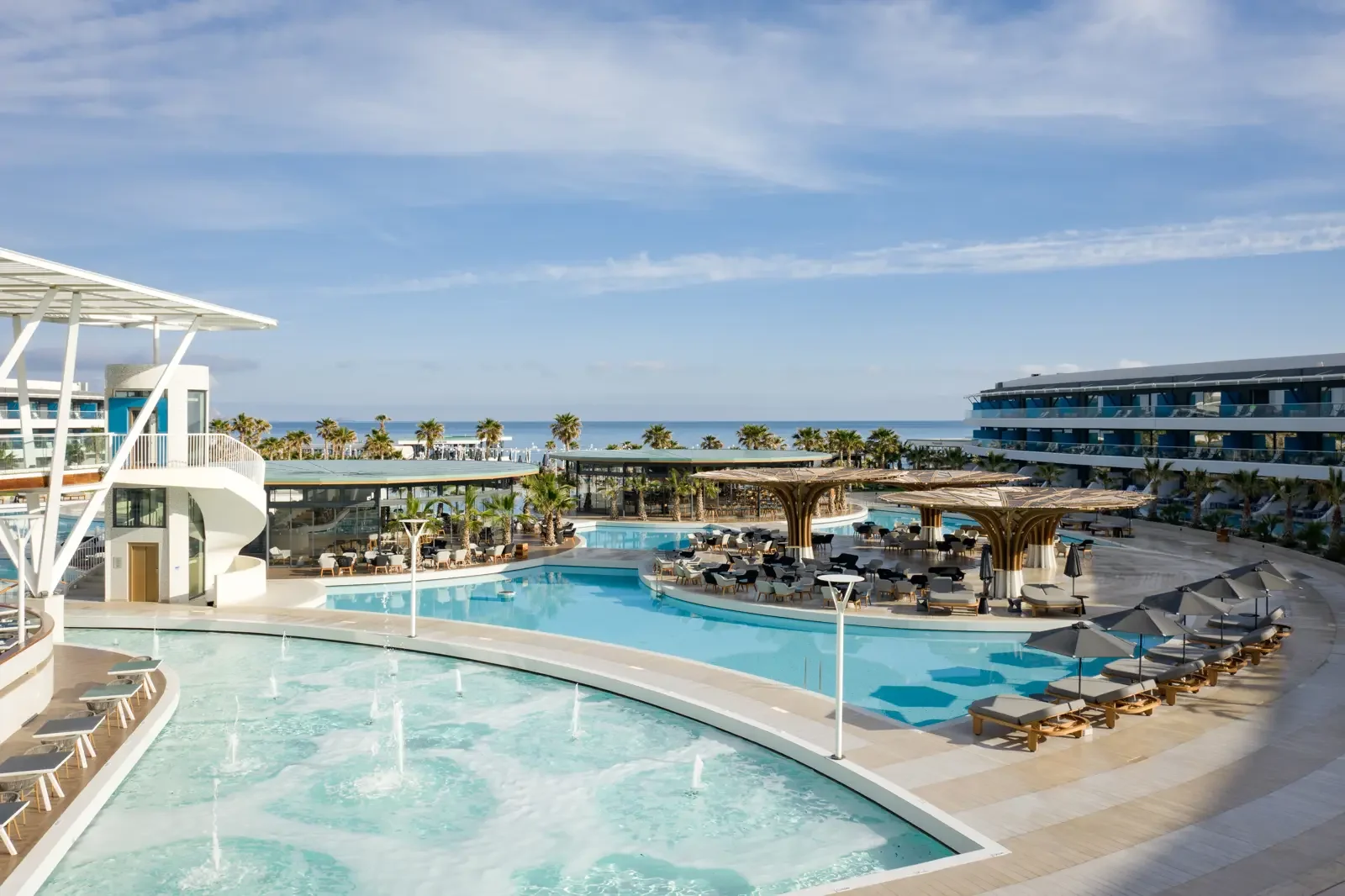 Zwembadgebied van een luxe resort met ligbedden, parasols en moderne architectuur, uitzicht op de oceaan en blauwe lucht.