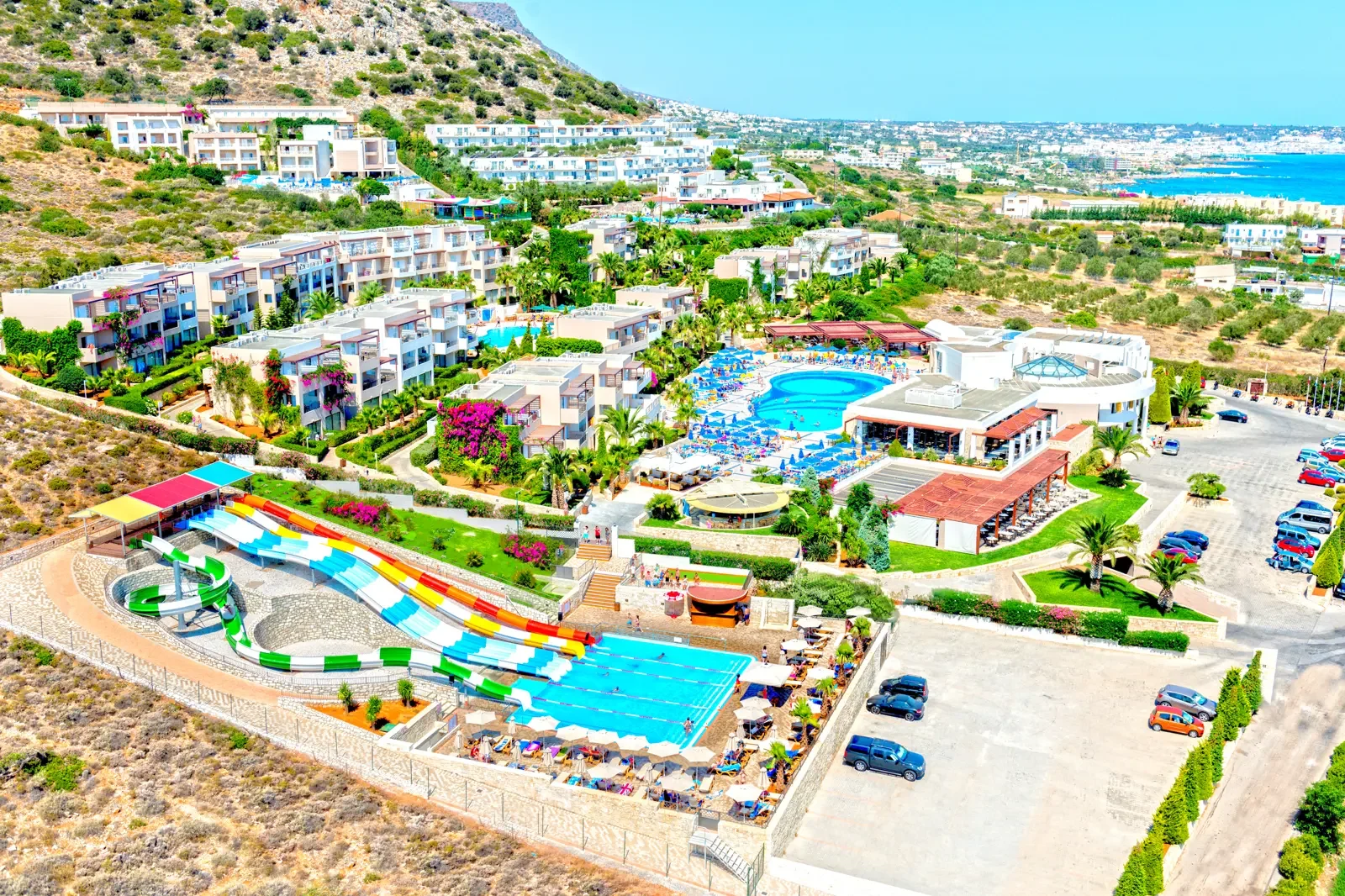 Luchtfoto van een luxe resort met meerdere zwembaden, glijbanen, moderne appartementen, tuinrijke omgeving en uitzicht op de zee en omliggende bergen.