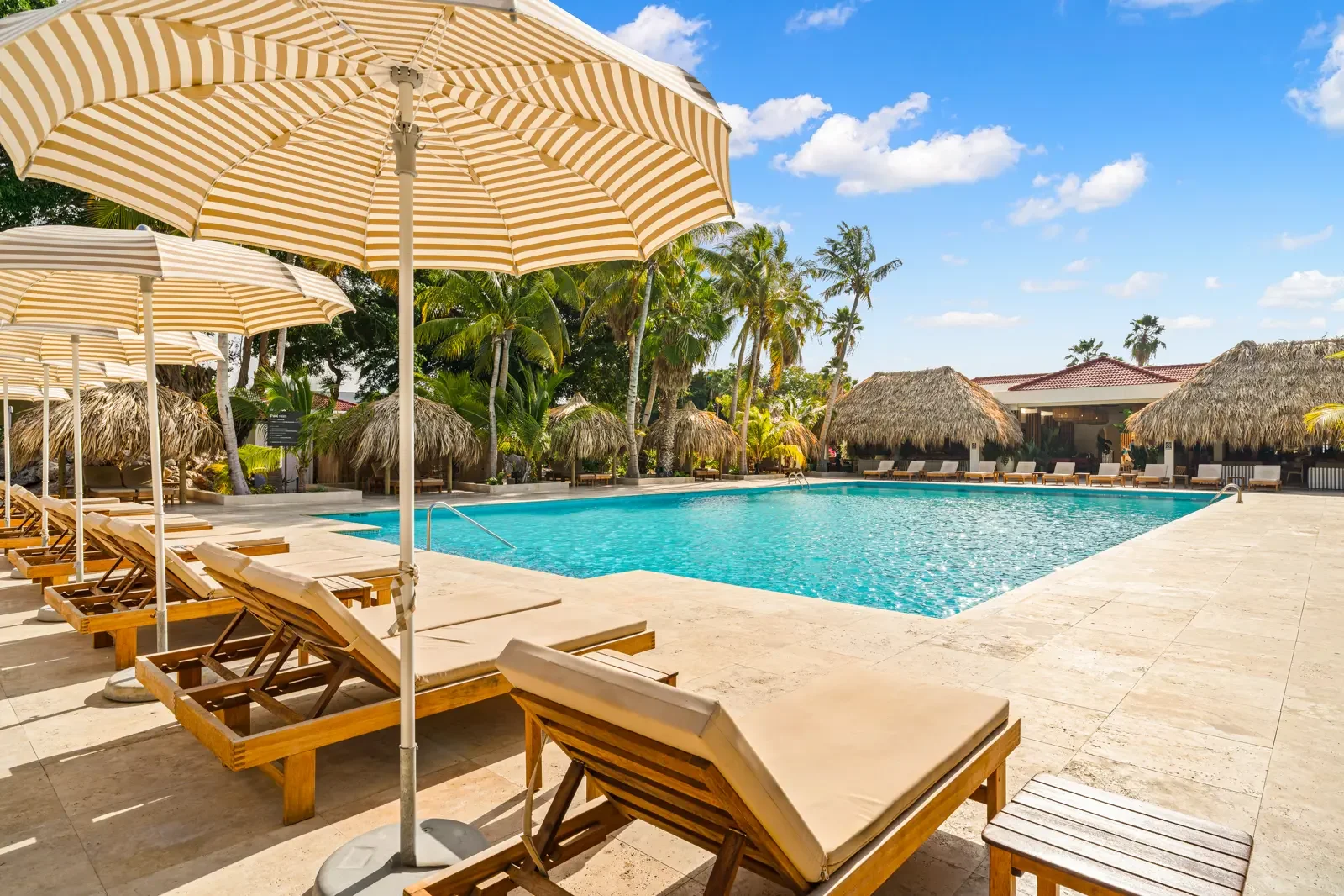 Zonovergoten zwembad met ligbedden en parasols, omgeven door palmbomen en thatagedeelten, in een tropisch resort onder een blauwe hemel met enkele wolken.