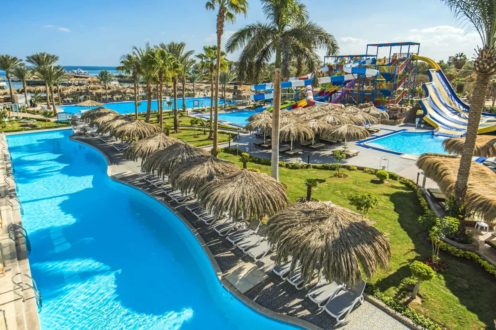 Zonder mensen, een zwembad met een waterspeelplaats met glijbanen, hangmatten onder rieten parasols, palm bomen, blauwe lucht en de zee op de achtergrond.