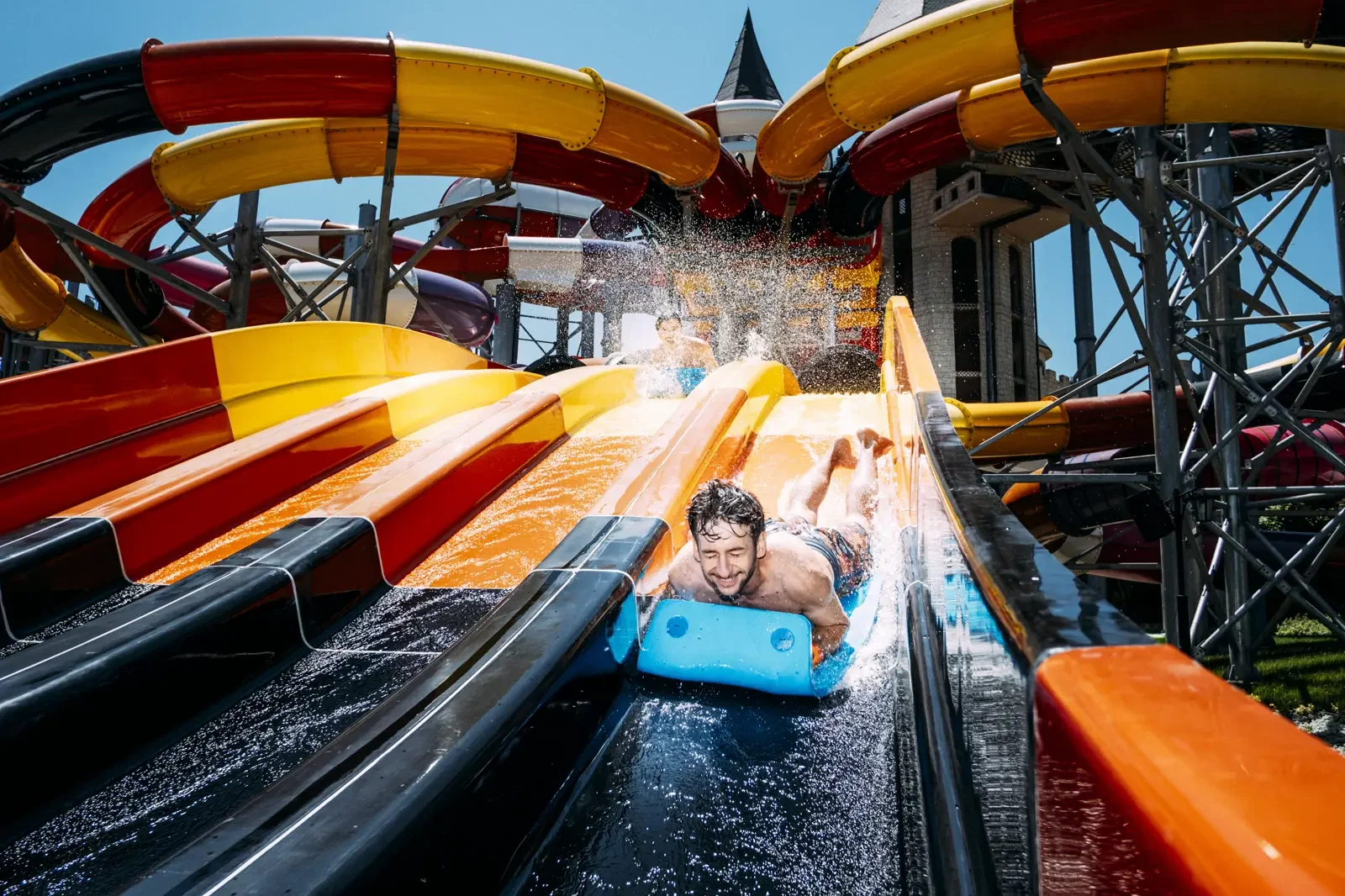 Mensen genieten van een waterglijbaan in een pretpark, waarbij ze afdalen met water over de glijbaan onder een heldere blauwe hemel.