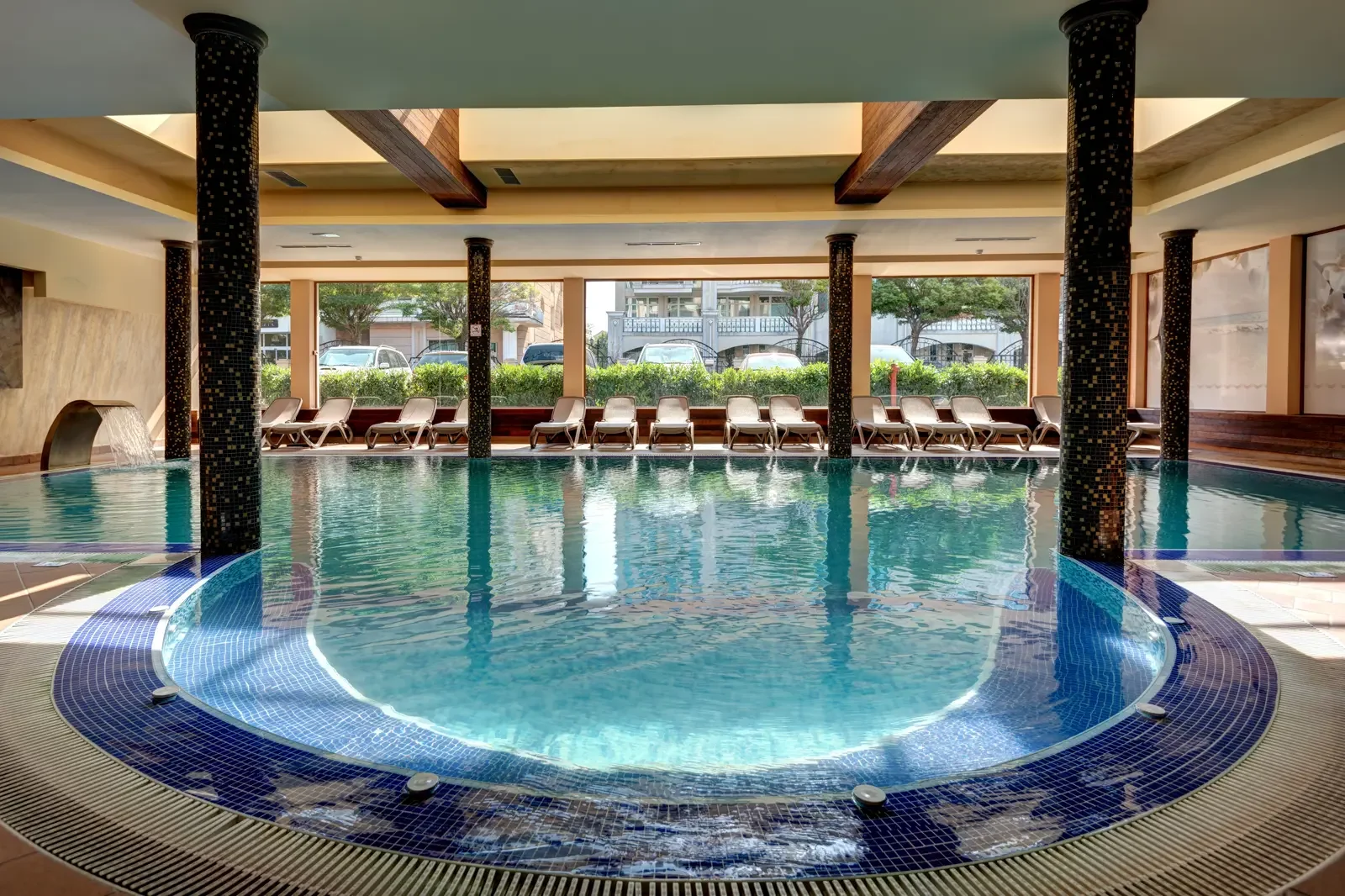 Binnenzwembad met ligstoelen langs het raam, grote ramen met uitzicht op een straat met auto's en huizen, enkele zwarte stenen versieringen, water dat uit een fonteintje stroomt.