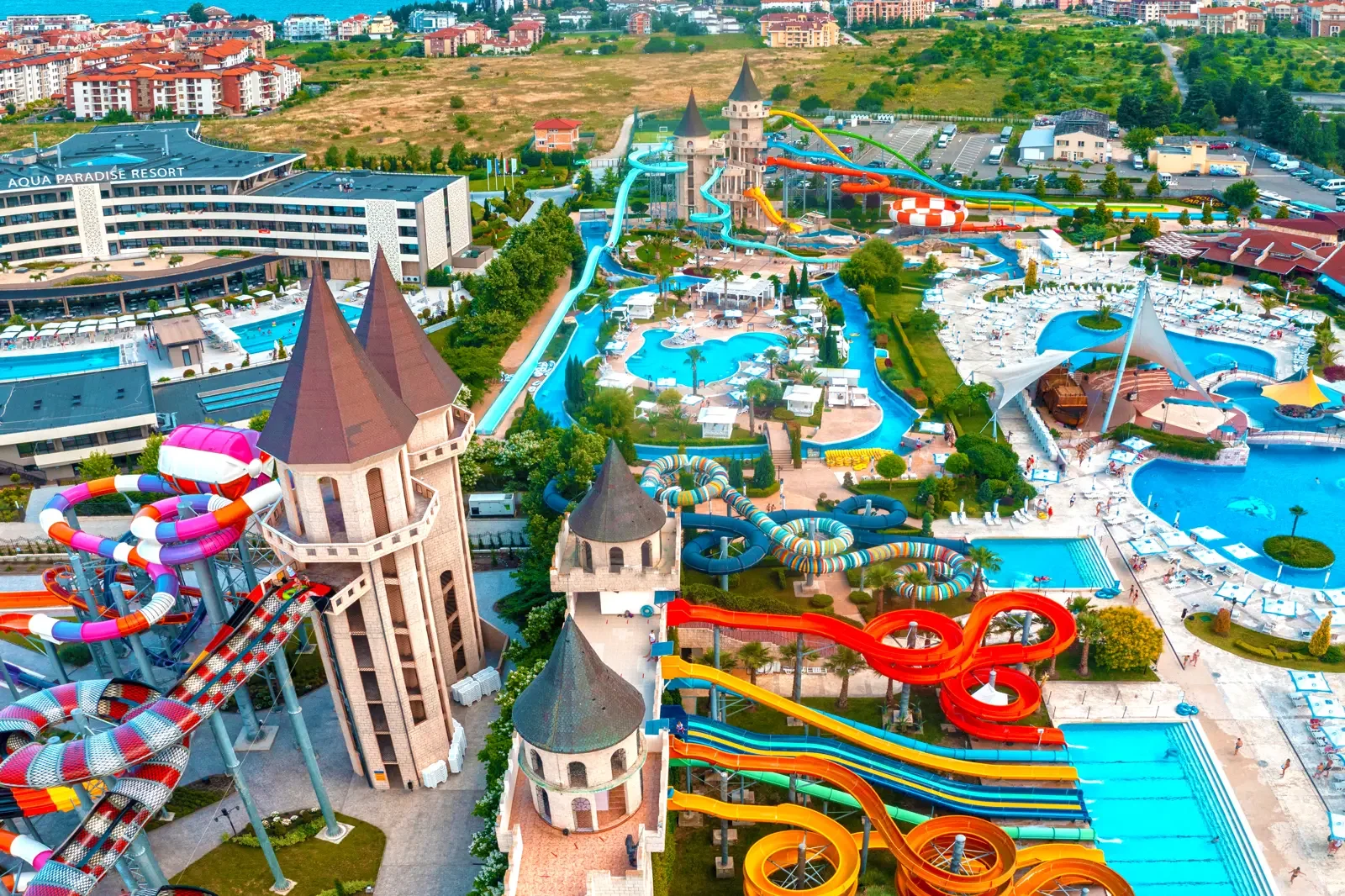 Luchtfoto van een waterpark met glijbanen, zwembaden en een kasteelachtig gebouw, omringd door gebouwen en groen landschap.