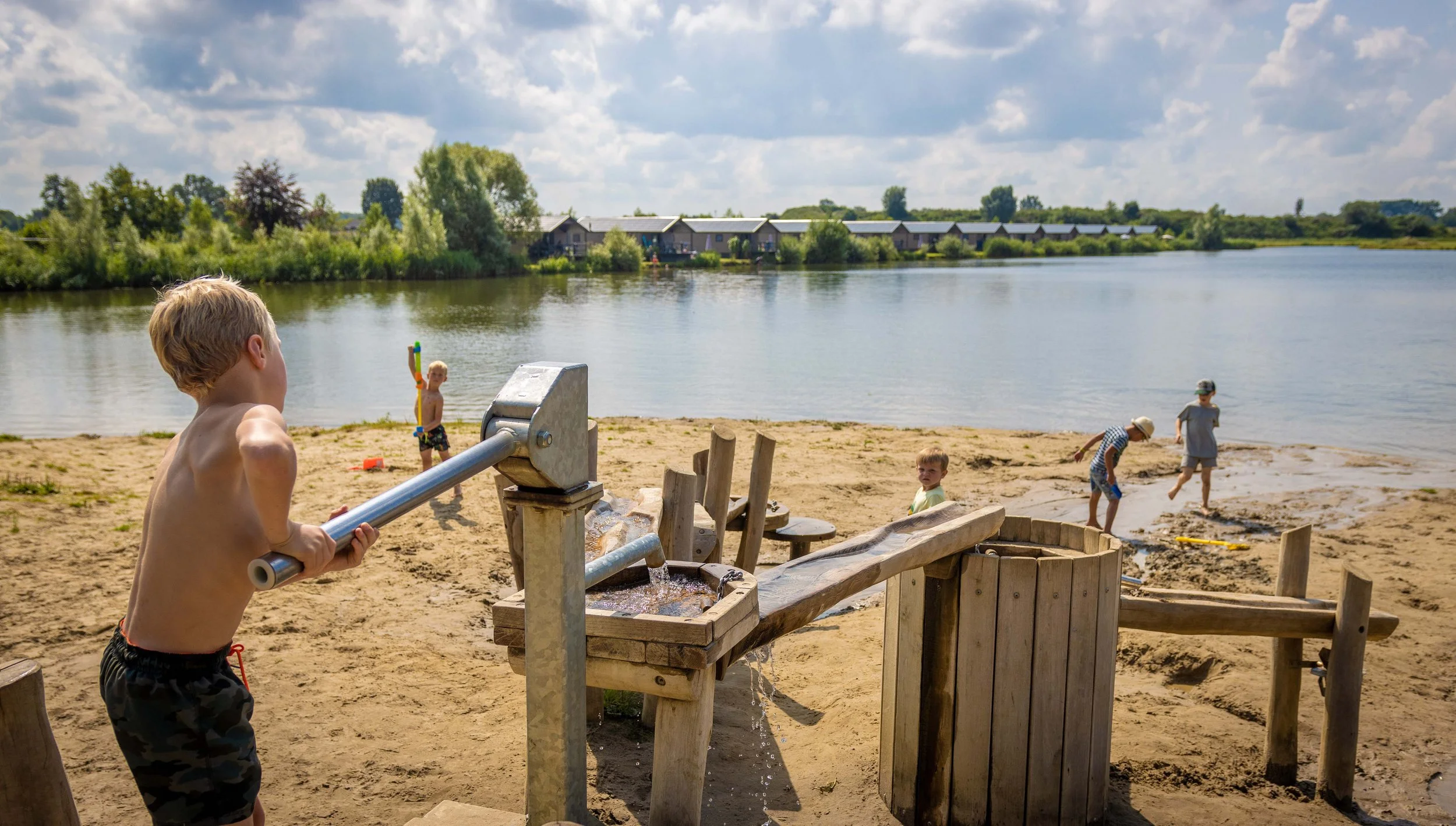 Kinderen spelen bij een watertappunt op een strand, met water dat uit een buis stroomt, langs een rivier met huizen op de achtergrond onder een bewolkte hemel.