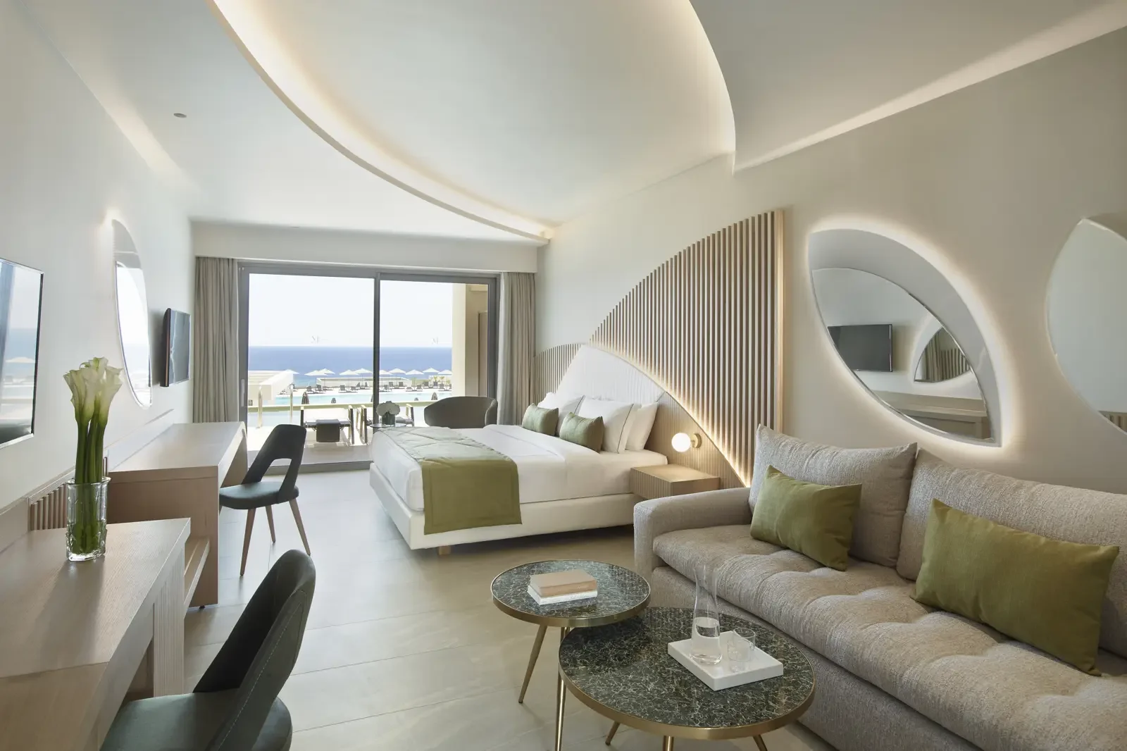 Luxe hotelkamer met groot bed, balkon met uitzicht op de zee, zithoek met bank en tafels, decoratieve muurreliëfs, planten en moderne meubels.