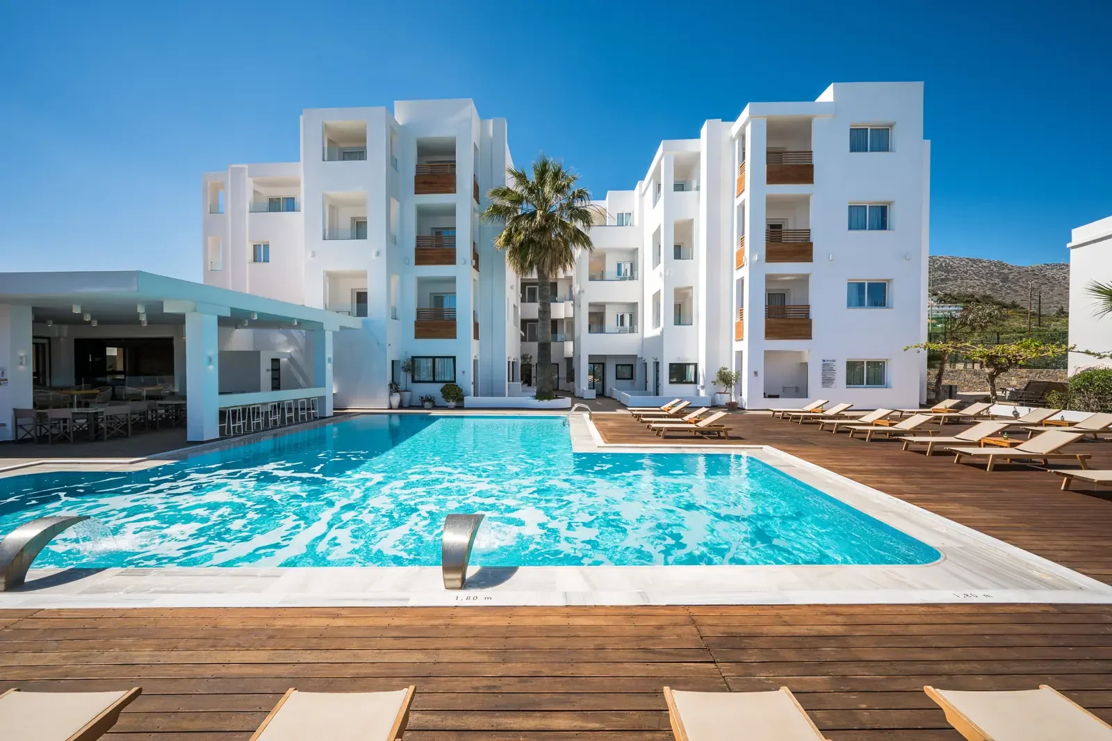 Zwembad met ligstoelen en een wit appartementencomplex met balkons op de achtergrond, palmbomen en een blauwe lucht.