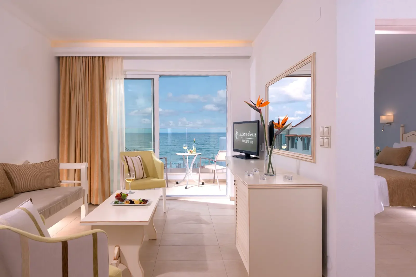 Een hotelkamer met uitzicht op de oceaan, voorzien van zit- en eetgedeelte, een tv, een balkon met een tafel en stoelen, en een aangrenzende slaapkamer.