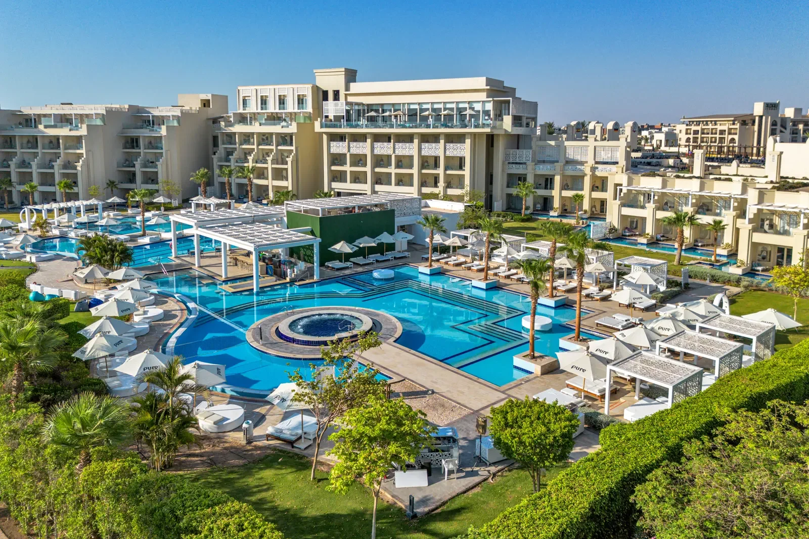 Luxe resort met groot zwembad, ligbedden, parasols en palmbomen, omgeven door moderne witte gebouwen.