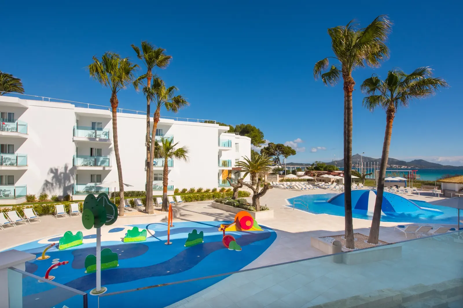 Zonnig zwembadcomplex met peuterbad en glijbanen, omgeven door palmbomen en ligstoelen, met een wit hotelgebouw en uitzicht op de zee in de achtergrond.