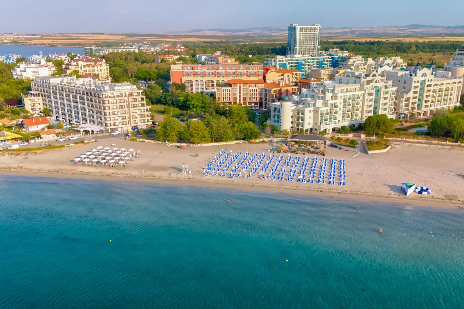 Luchtfoto van een vakantieoord met appartementen en hotels aan een strand met ligbedden en parasols, en een helderblauwe zee op de voorgrond