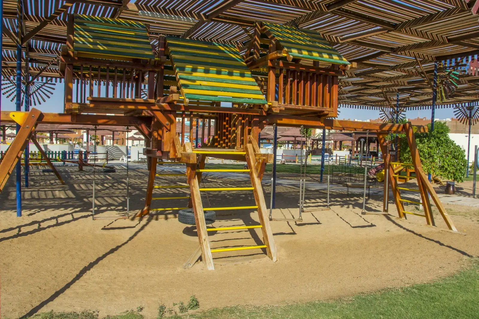 Speeltoestellen en schommels op een kinderspeelplaats, onder een grote structuur met houten en kleurrijke onderdelen.