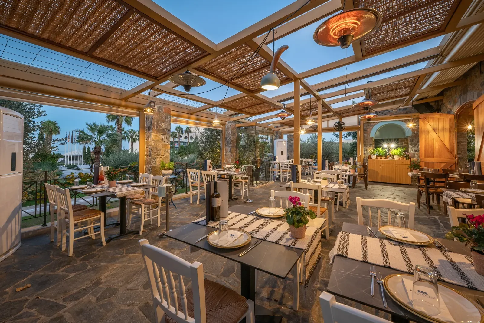 Een gezellige, open-air restaurant met wit en houten stoelen, tafels gedekt met servies en planten, omgeven door palmbomen en terraslampen, binnen een rustiek gebouw met een rieten dak en stenen muur.