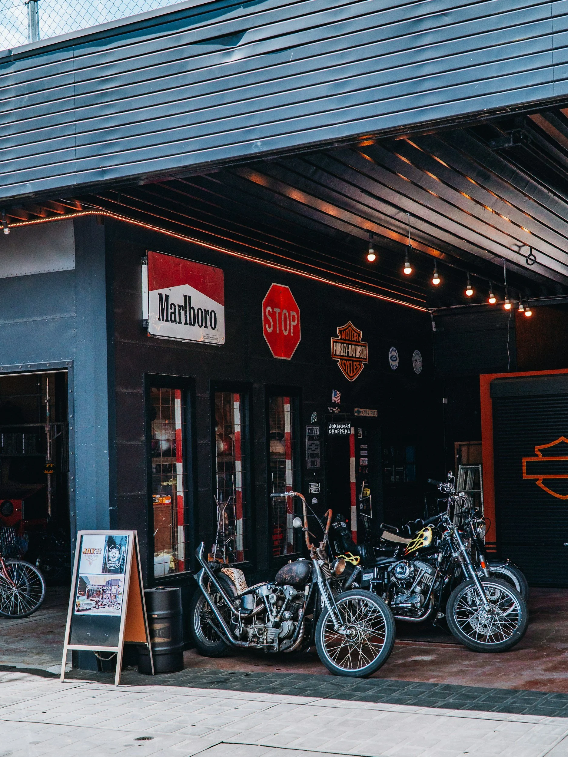 Exterior of a motorcycle shop with motorcycles parked outside, Harley Davidson logo on the wall, and a signboard displaying motorcycle illustrations and information.