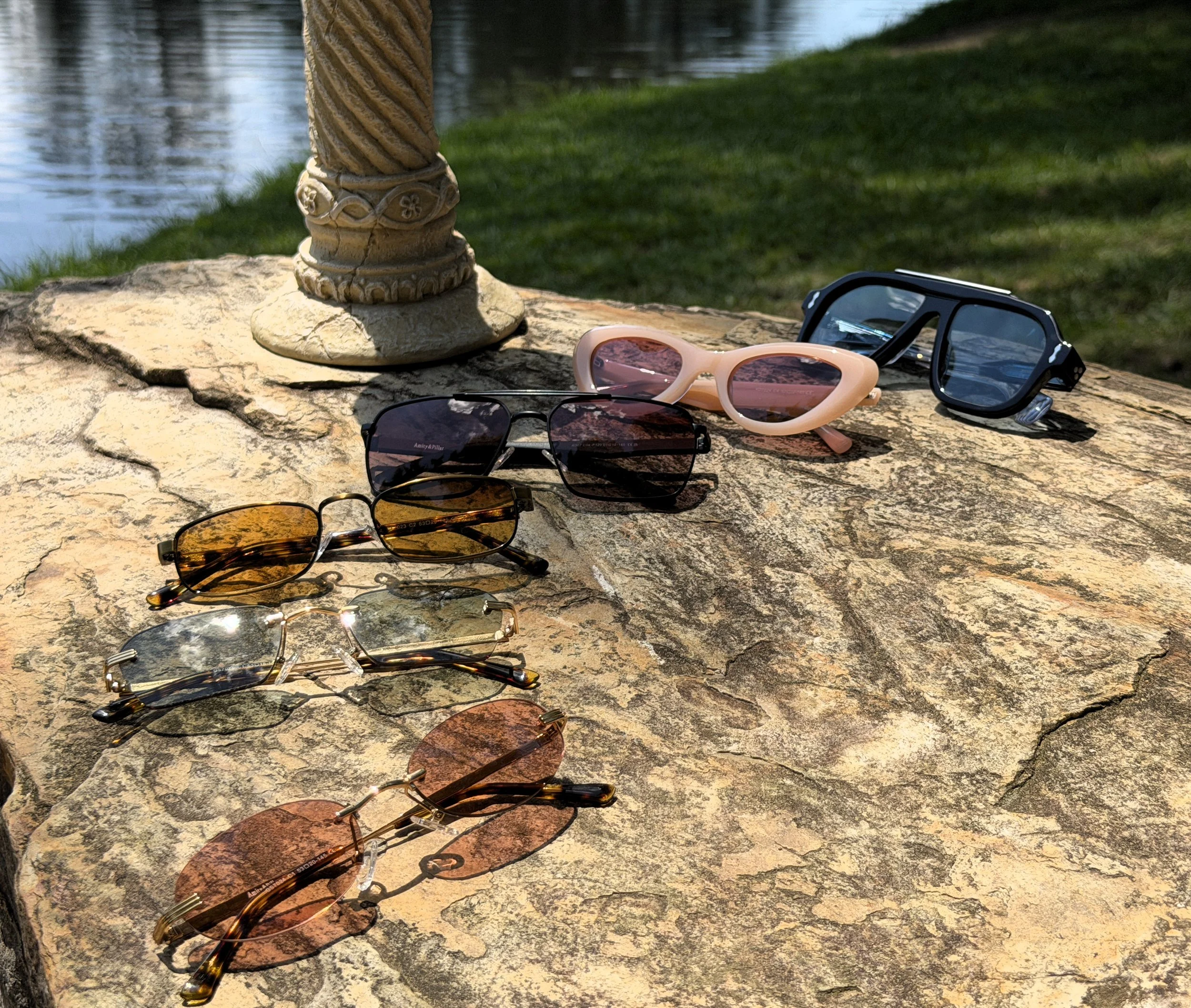 Several pairs of sunglasses are laid out on a large, flat rock near a body of water with a grassy area in the background, and a carved stone column or sculpture also resting on the rock.