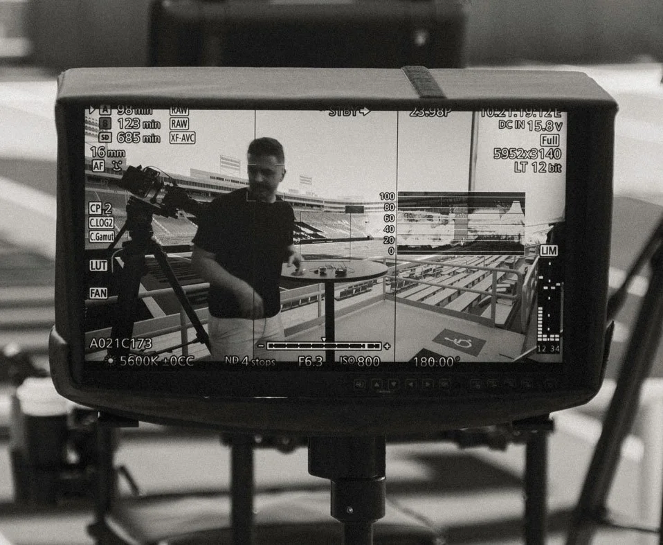 A person standing next to a round table on a stadium seating area, seen through a camera monitor, with camera settings displayed on the screen.