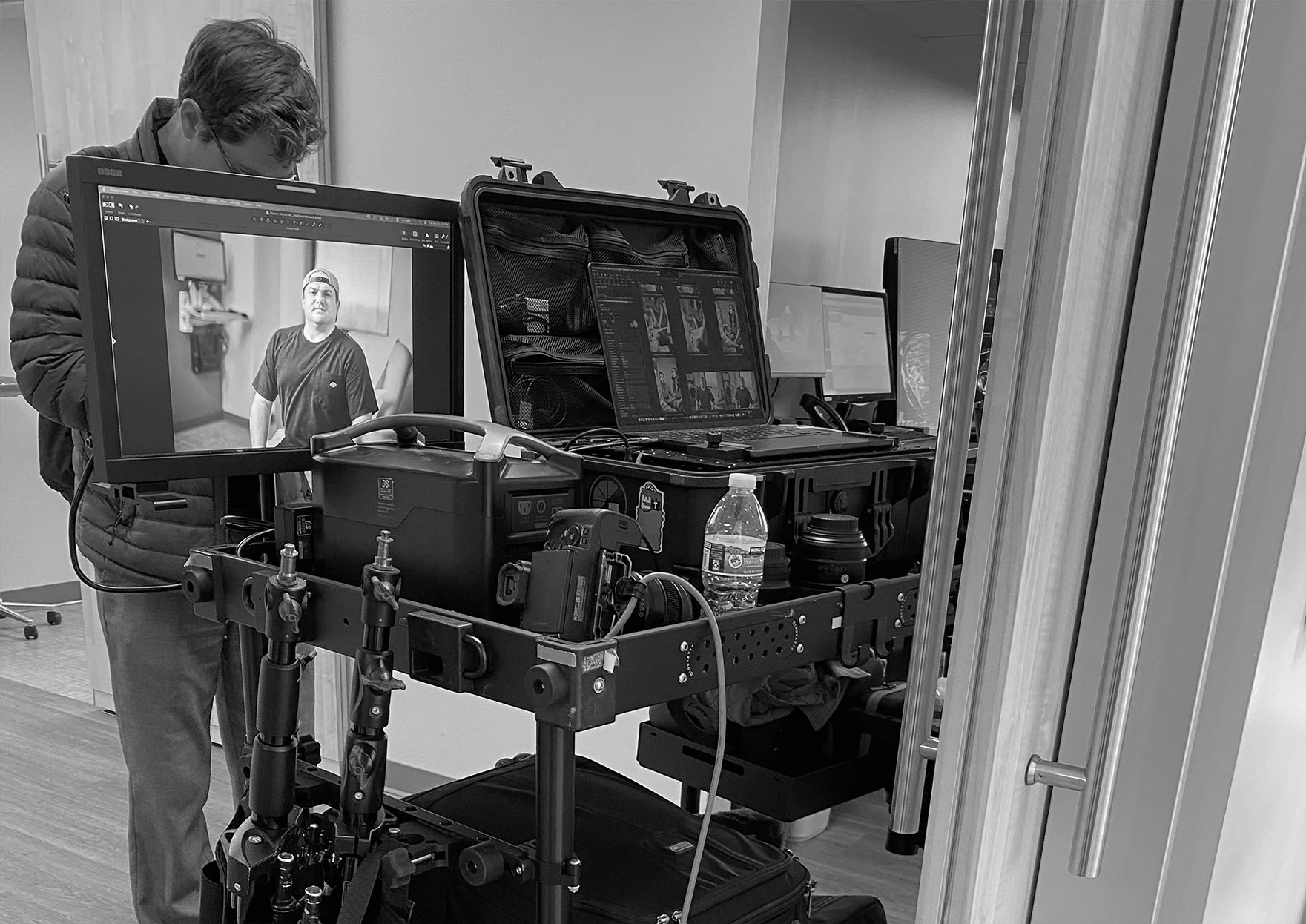 Black and white photo of a man with glasses in a photography or video studio, working with camera equipment, monitors, and a water bottle on a wheeled cart.