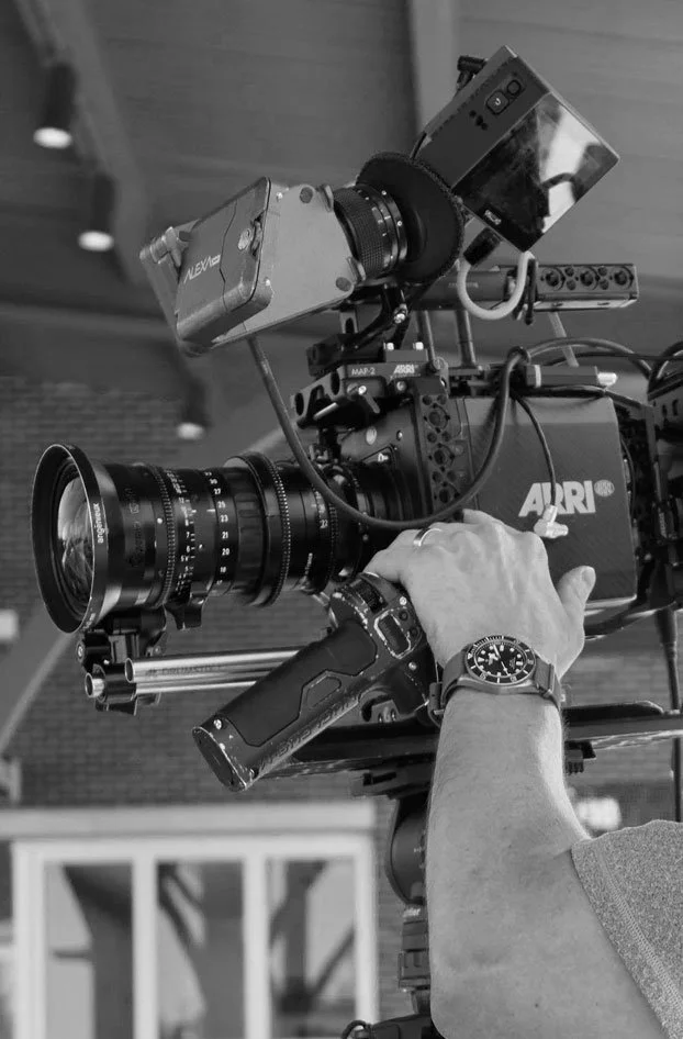 Black and white photo of a professional video camera with a person's hand adjusting it. The person is wearing a wristwatch.