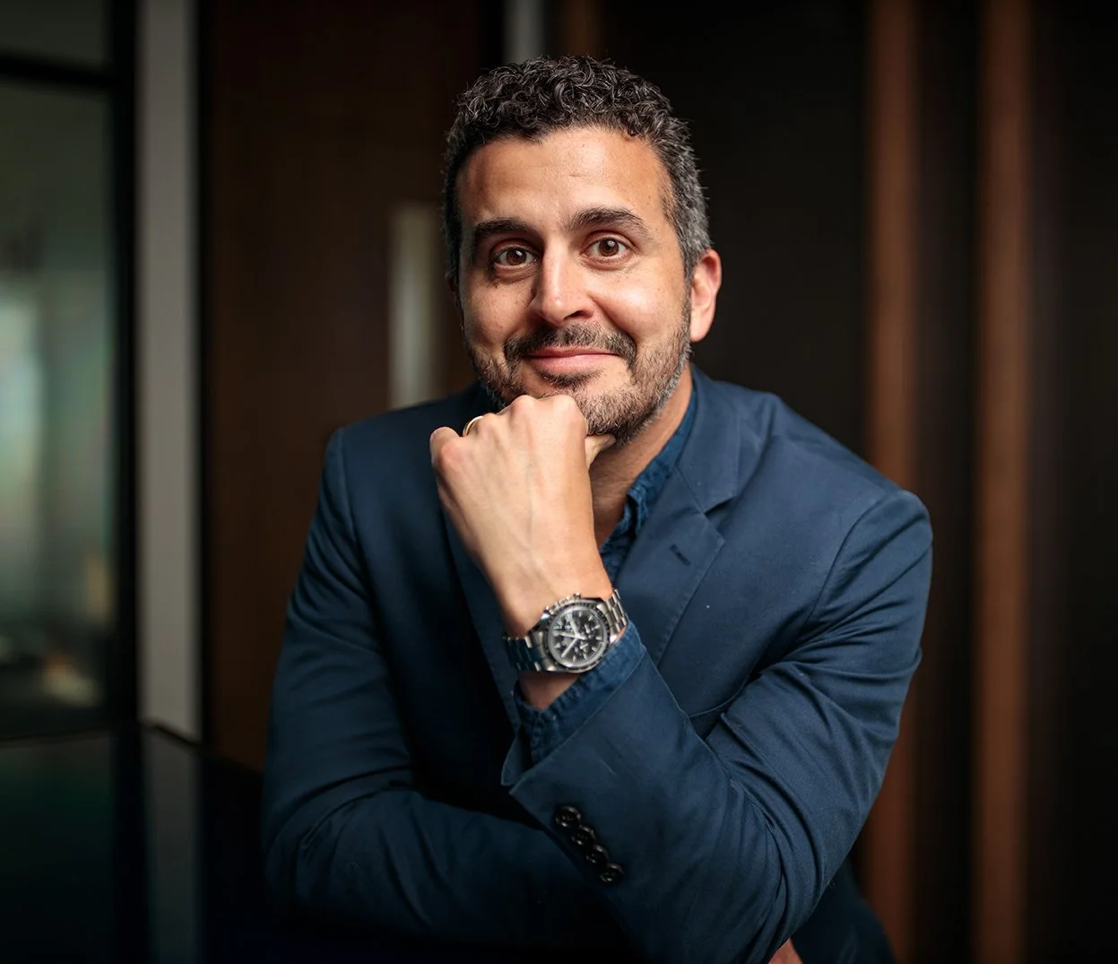 A man with short curly hair and a beard, wearing a blue blazer and watch, sitting with his chin resting on his hand, smiling at the camera.