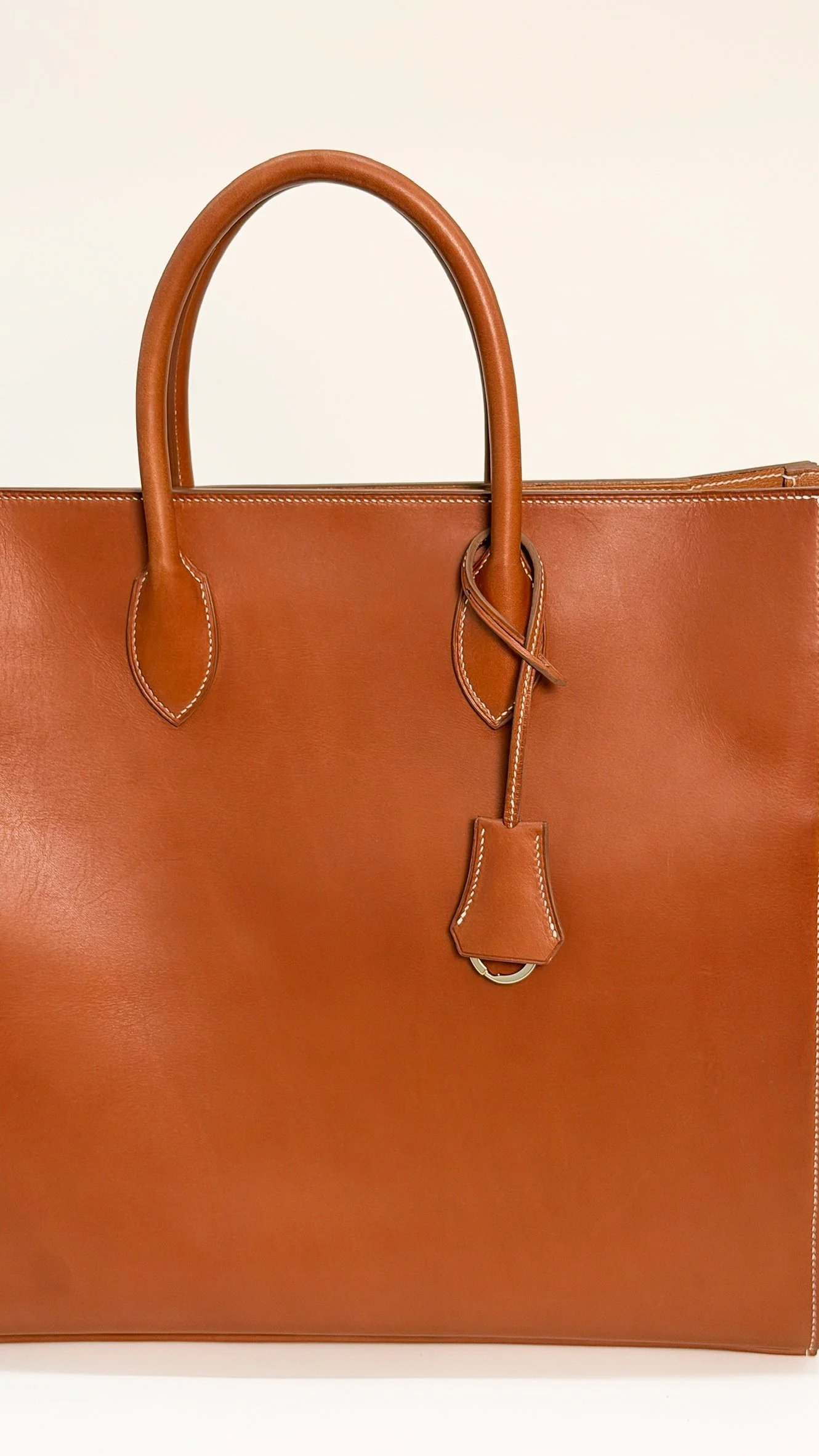 A close-up of a brown leather handbag with two handles, white stitching, a hanging leather tag, and a gold ring.