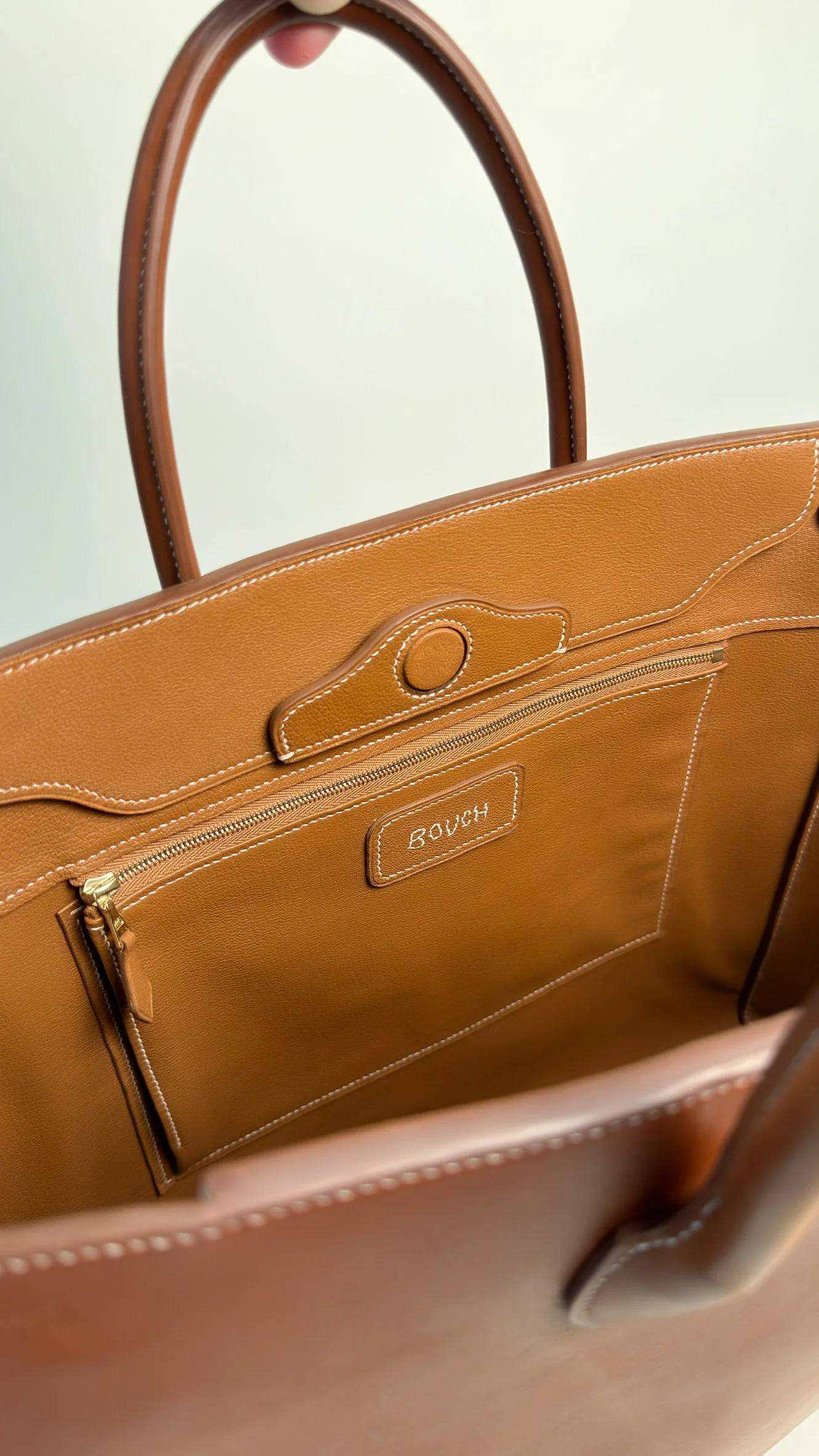 A tan leather handbag with a handle, featuring a front zippered pocket and a label that reads 'BOUCH' inside the bag, with white stitching details.