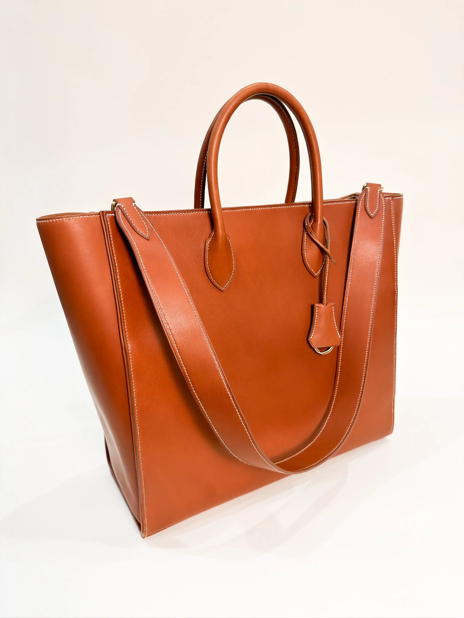 A large, brown leather tote bag with two handles and decorative stitching, set against a plain background.