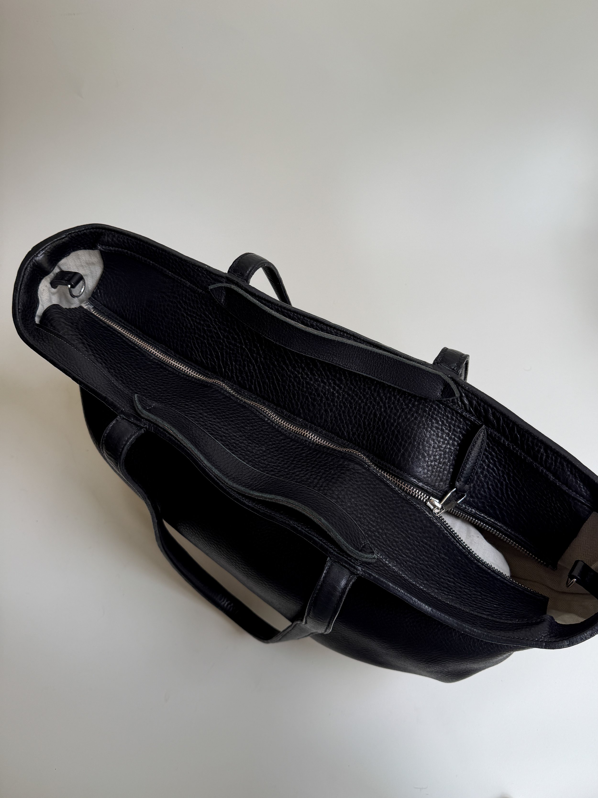 Top view of a black textured leather handbag with a zipper and handle, resting on a white surface.