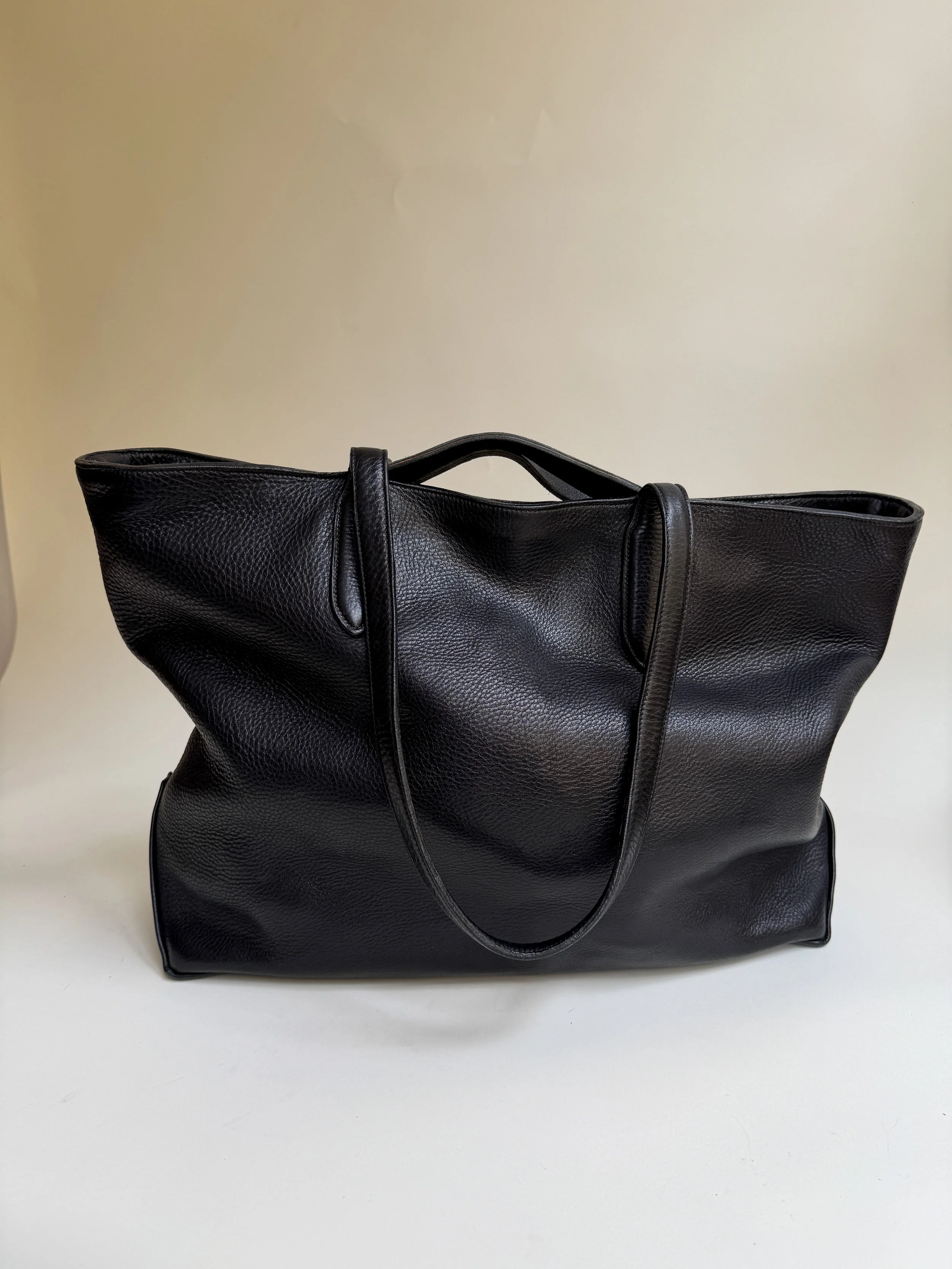 A black leather tote bag with two handles on a white surface against a beige wall background.