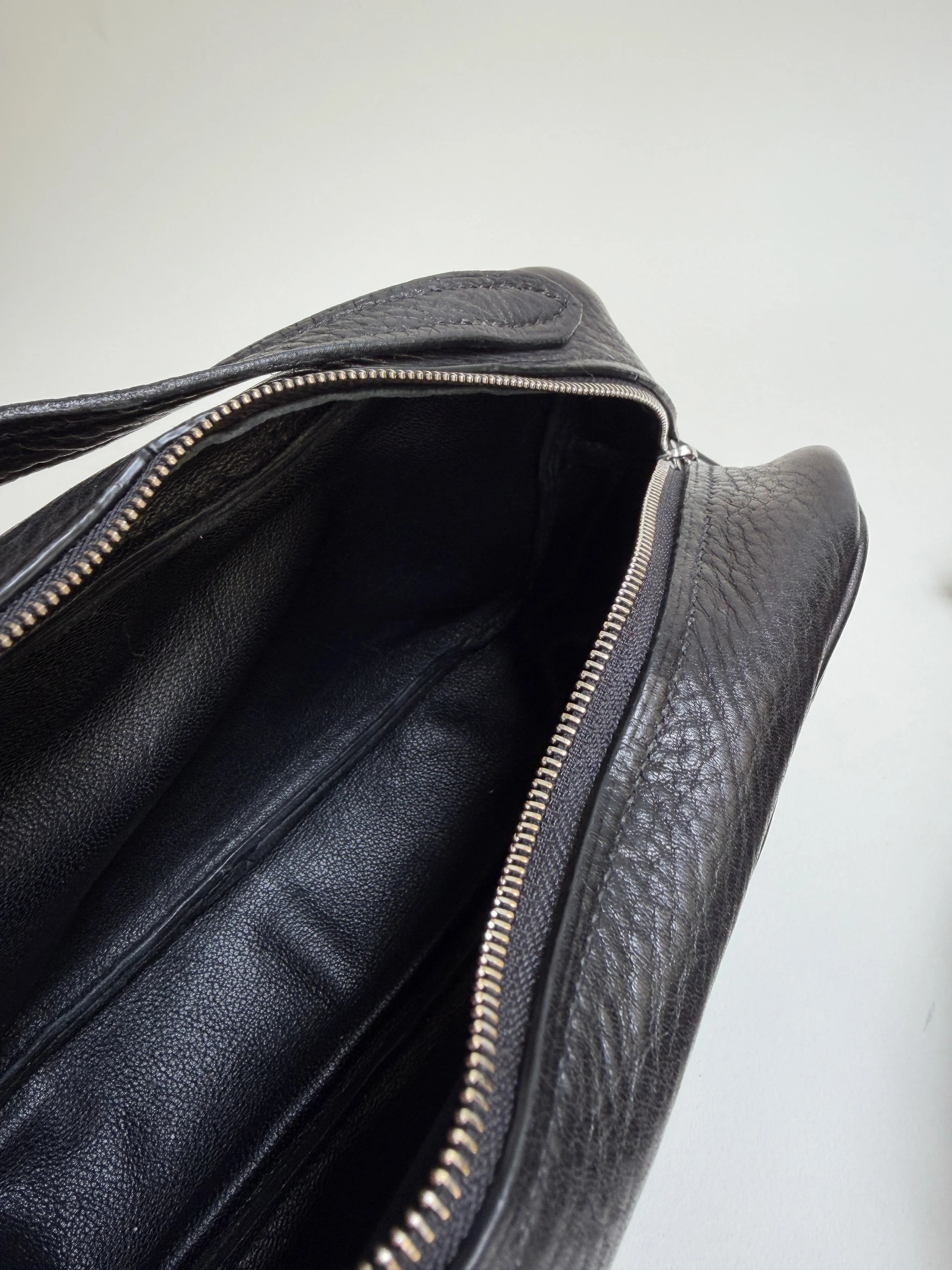 Close-up of a black leather bag with an open zipper, revealing a dark interior, against a plain white background.