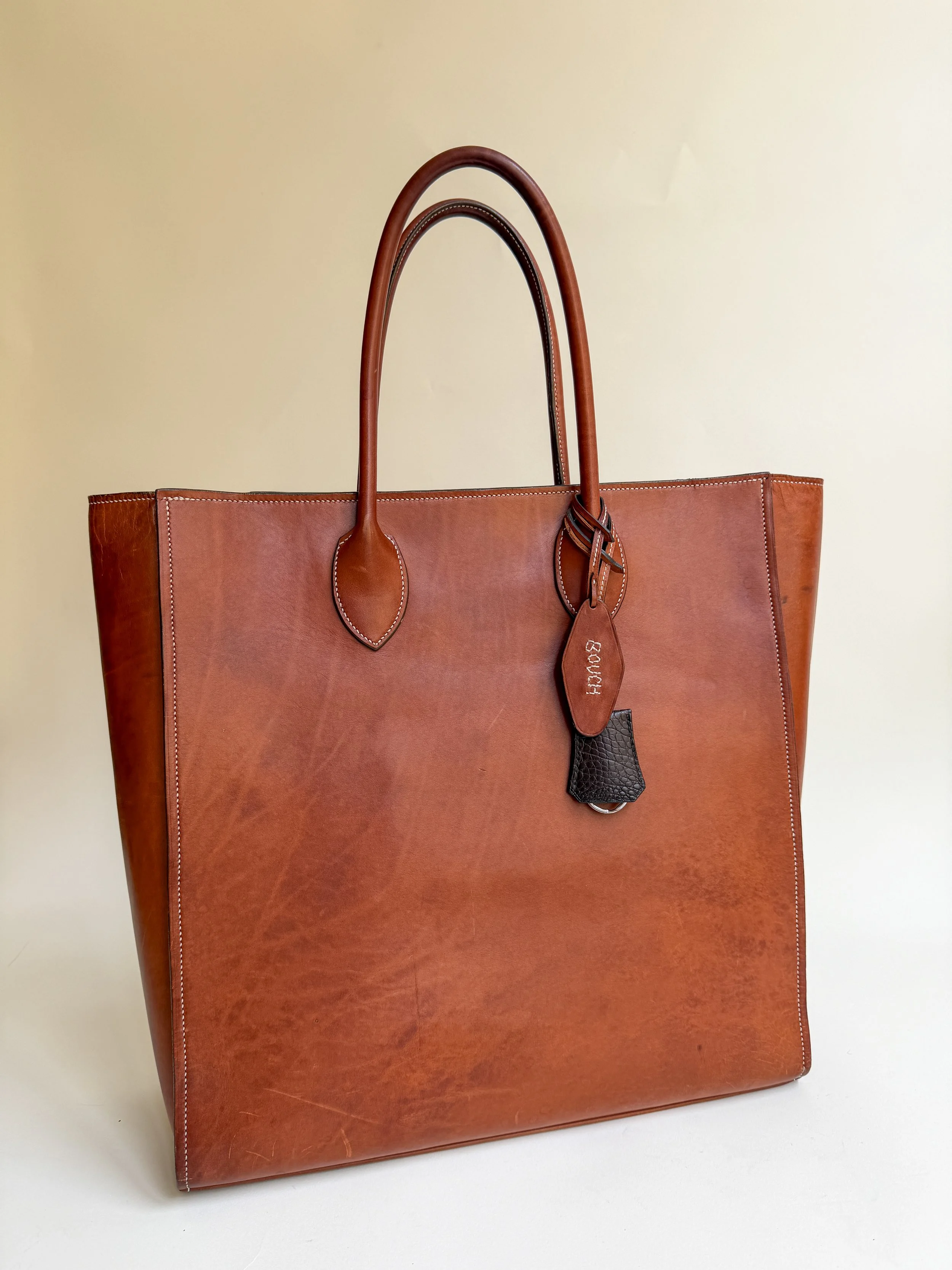 A large brown leather tote bag with two handles and a tag hanging from one handle, positioned against a beige background.
