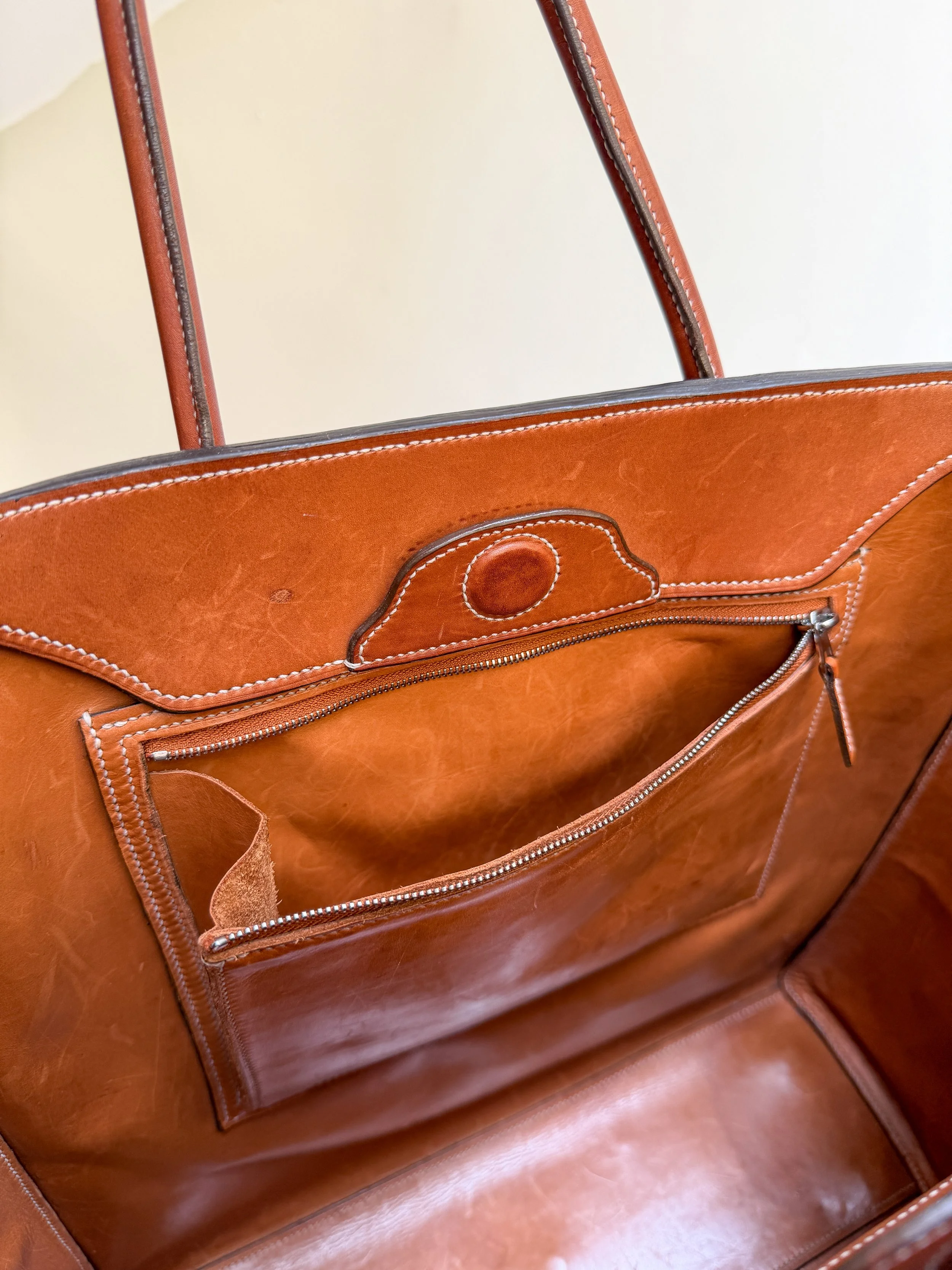 A close-up of a brown leather handbag with a front zipper pocket, white stitching, and two handles, on a wooden surface