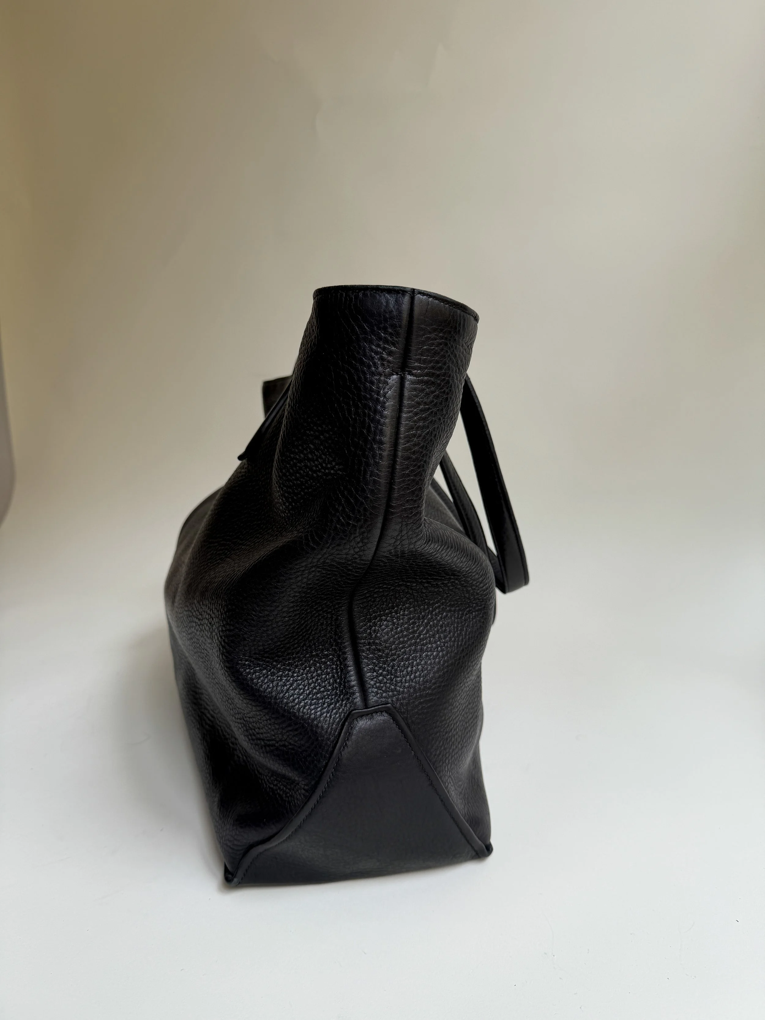 Black leather handbag with textured surface, standing upright on a white surface, against a plain light-colored background.