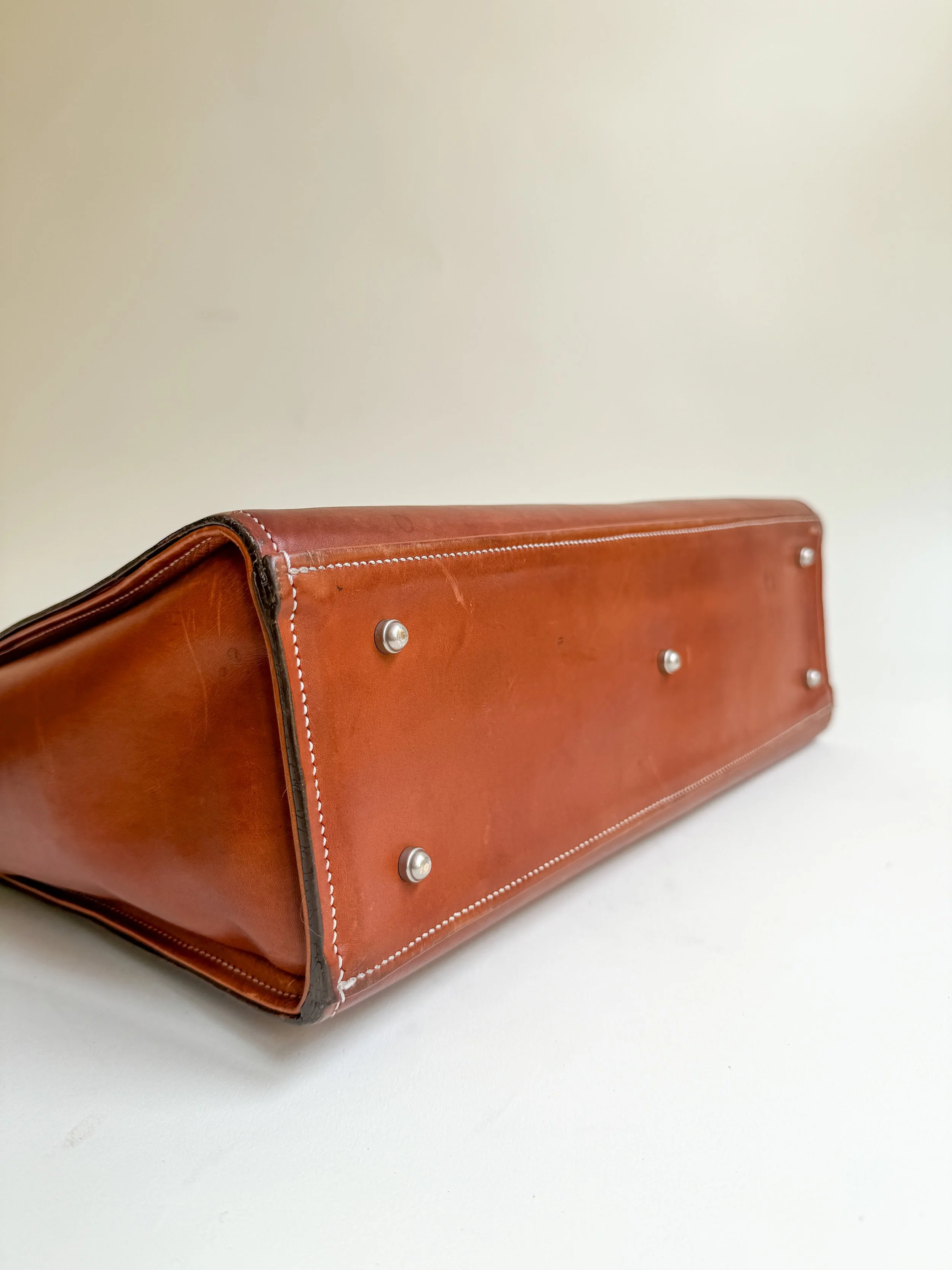 Close-up of the bottom corner of a brown leather briefcase with metal feet, white stitching, and zippers, placed on a white surface against a plain white wall.