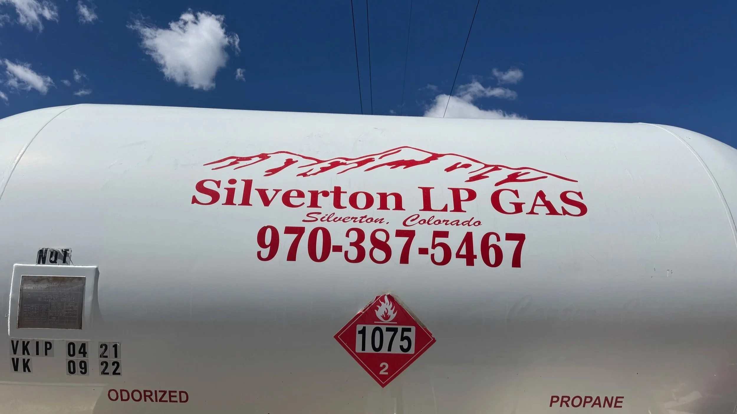 A white propane tank with red text and mountain logo that reads 'Silverton LP Gas, Silverton, Colorado, 970-387-5467.' The tank has various labels, including a diamond warning label with a flame symbol and '1075.' The sky is blue with a few white clouds.