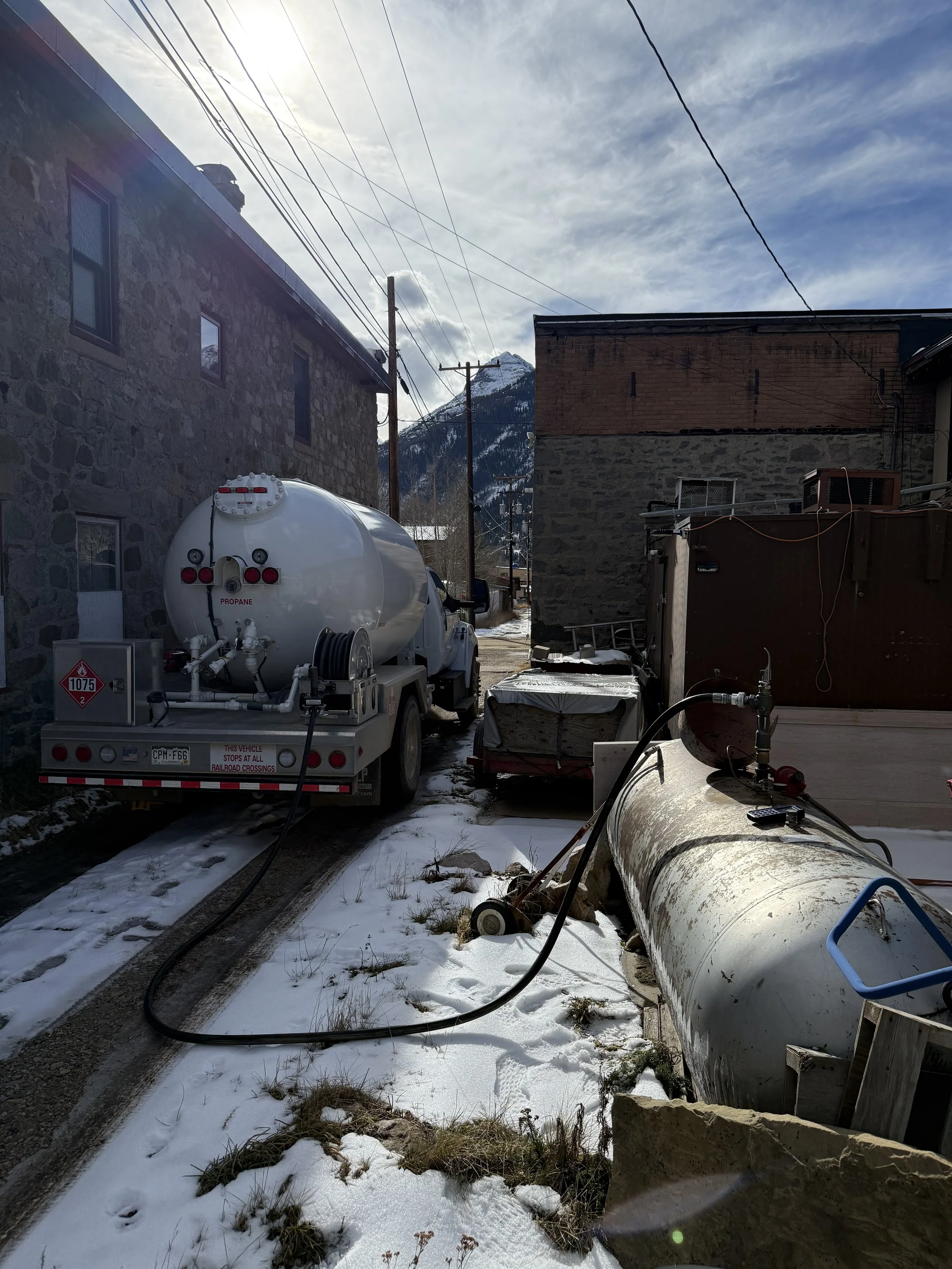 Snow-covered alleyway with a propane tank on a trailer and large pipes on the ground, between two buildings, with mountains and cloudy sky in the background.