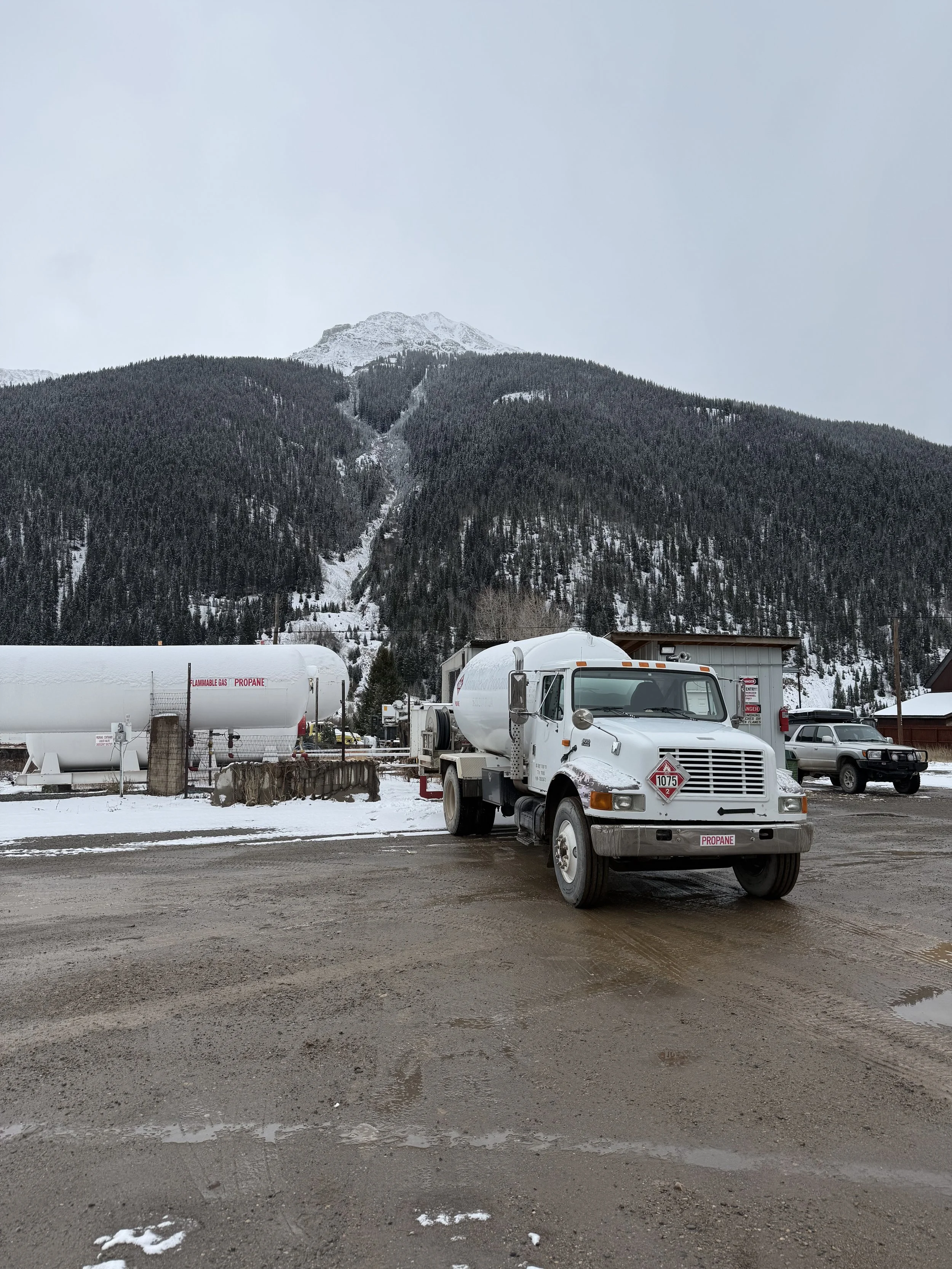 A snow-covered parking lot with a propane gas truck and an industrial vehicle, set against a backdrop of pine trees and snow-capped mountains under a cloudy sky.
