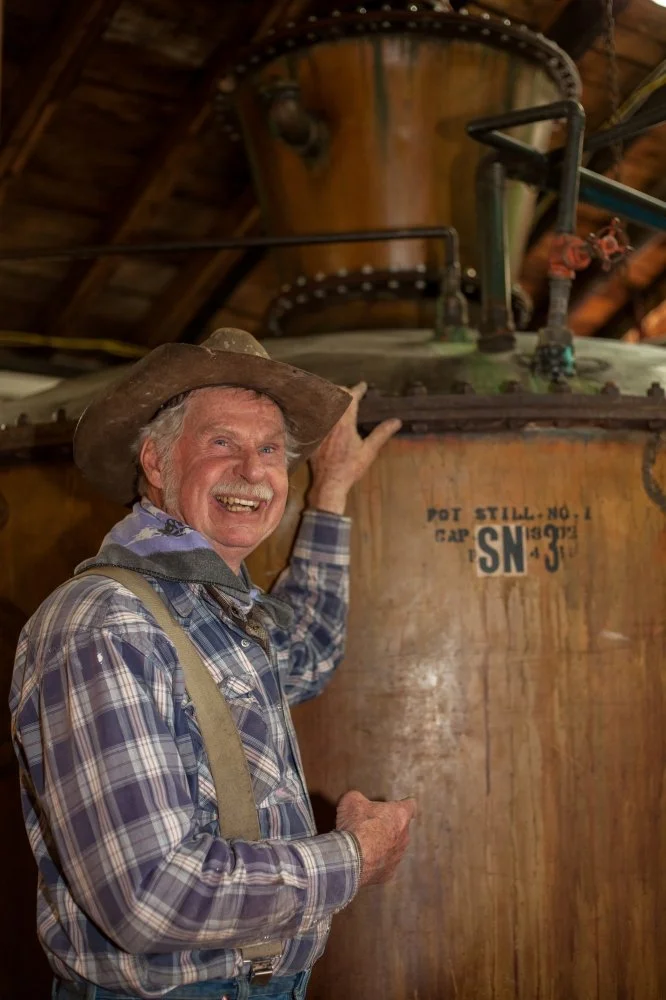 An elderly man in plaid shirt, suspenders, and cowboy hat smiling and pointing at a large, weathered industrial tank inside a rustic wooden building.