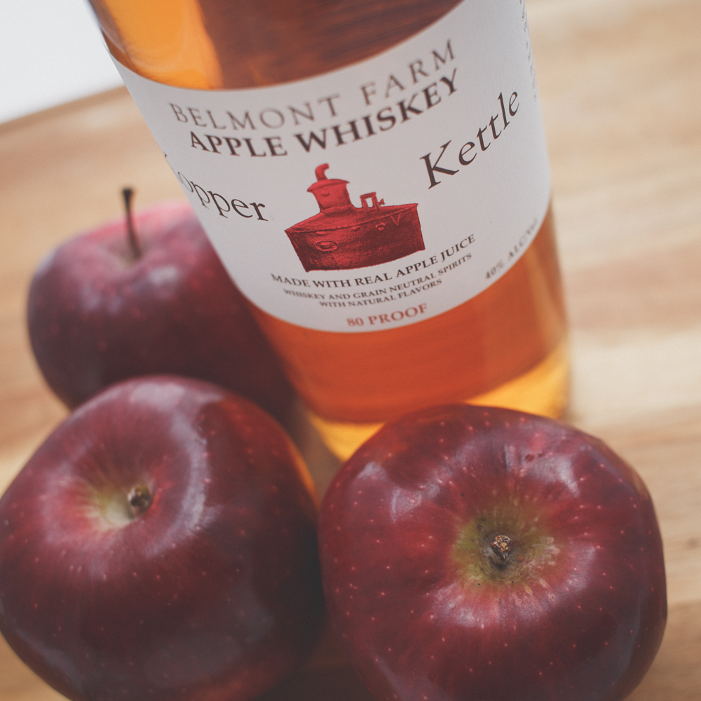 A bottle of Belmont Farm apple whiskey and three red apples on a wooden surface.
