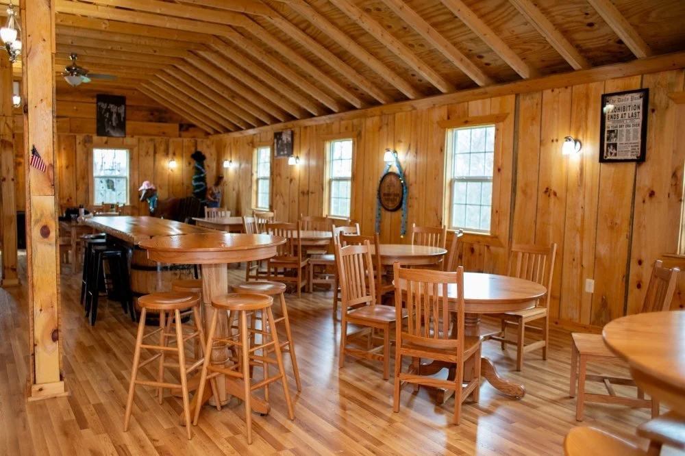 Interior of a cozy wooden restaurant or café with round tables, wooden chairs, a high bar table with stools, and warm lighting on wooden walls.