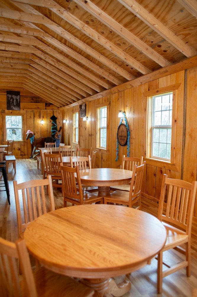 Interior of a rustic wooden dining room with round tables and chairs, wood-paneled walls, windows allowing natural light, and decorative wall hangings including a wreath and banners.