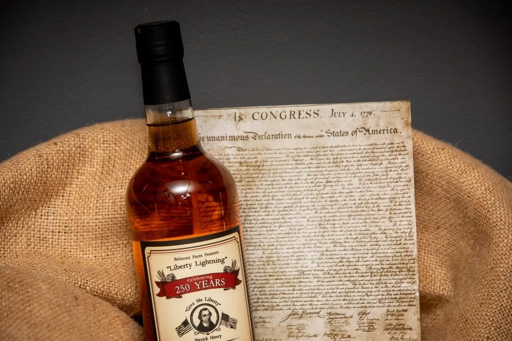 A bottle of whiskey labeled 'Liberty Lightning' with a 250-year celebration sticker, positioned in front of a historical document on burlap fabric.