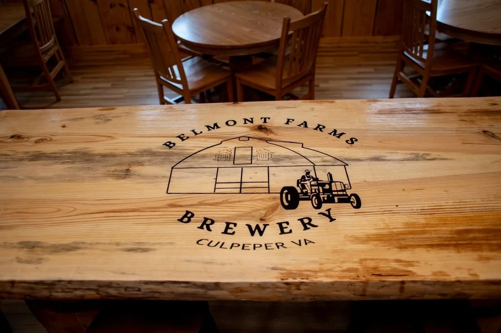 Wooden table with the logo of Belmont Farms Brewery and an illustration of a barn, a tractor, and a farmer, located in Culpeper, Virginia.