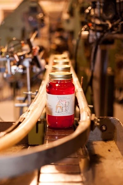 A red candle in a glass jar with a Christmas label, placed on an industrial conveyor belt in a factory setting.