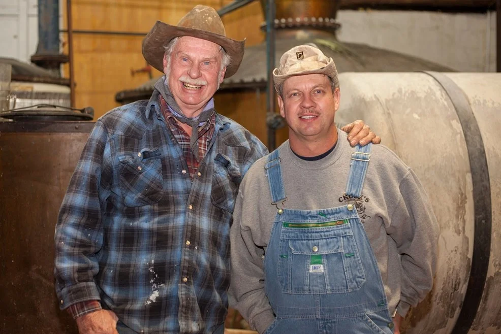 Two men in farm attire standing together indoors, smiling. The man on the left wears a big cowboy hat, plaid shirt, and has his hand on the shoulder of the man on the right, who wears a cap and overalls.