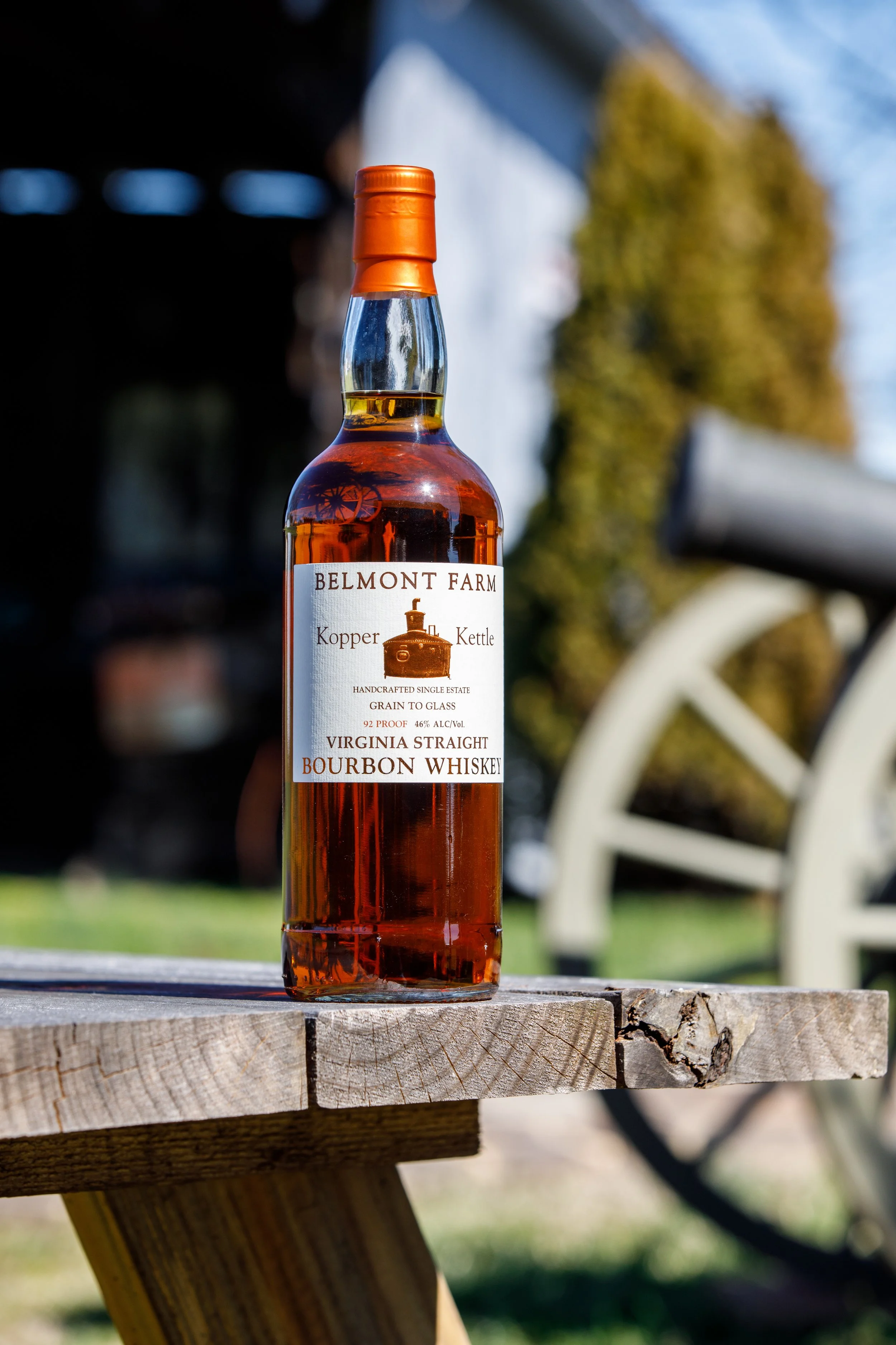 A bottle of Belmont Farm bourbon whiskey with a clear, amber liquid inside, placed on a wooden surface outdoors on a bright day, with a blurred background of trees and a white fence.