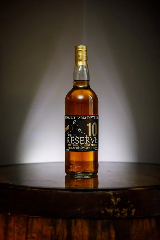 A bottle of Belmont Farm Distillery Special Reserve 10-year-old Virginia whiskey with amber color, placed on a wooden surface with a spotlight above, against a dark background.