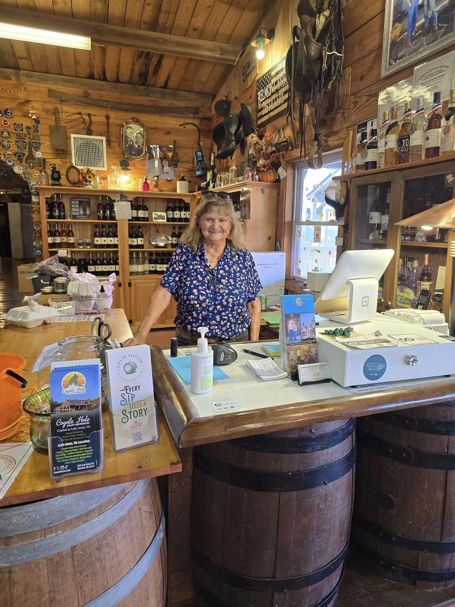 A smiling woman behind a wooden counter in a rustic store. The background has wooden walls decorated with vintage and western items, shelves with bottles, and a window with a view outside.