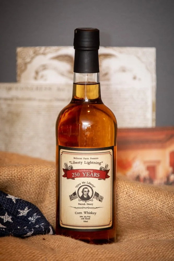 A bottle of corn whiskey with a label commemorating 250 years, featuring an image of Patrick Henry, placed on burlap fabric with background items including a paper and a small box.