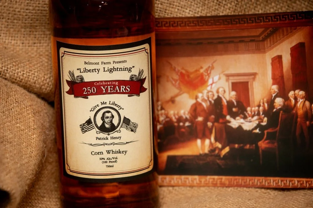 A bottle of Belmont Farm's Liberty Lightning corn whiskey celebrating 250 years, with a label featuring a portrait of Patrick Henry and American flags, placed next to a framed painting of the signing of the Declaration of Independence.