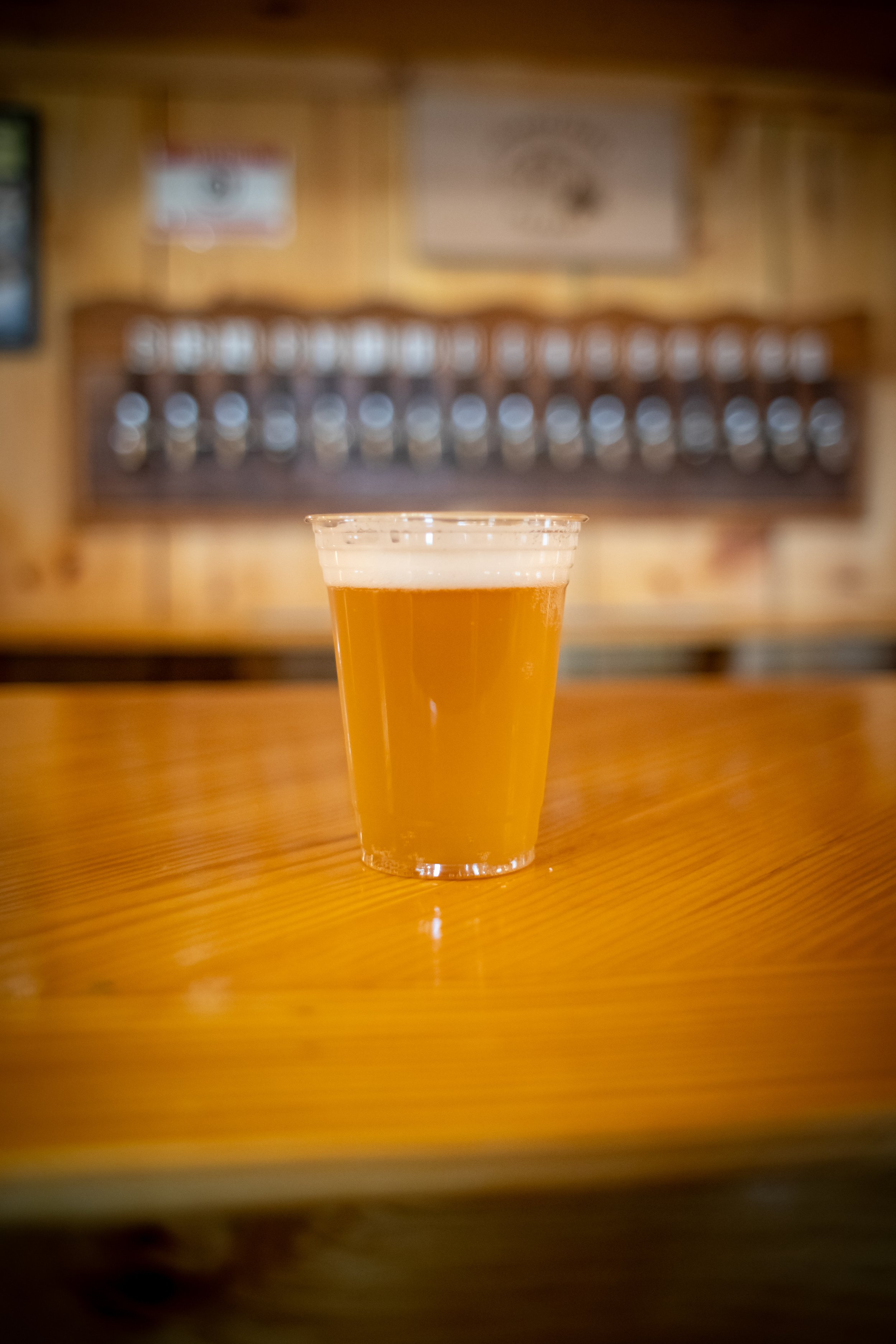 A clear plastic cup filled with a pale amber beer on a wooden surface with a blurred background of wooden walls and wall decorations.