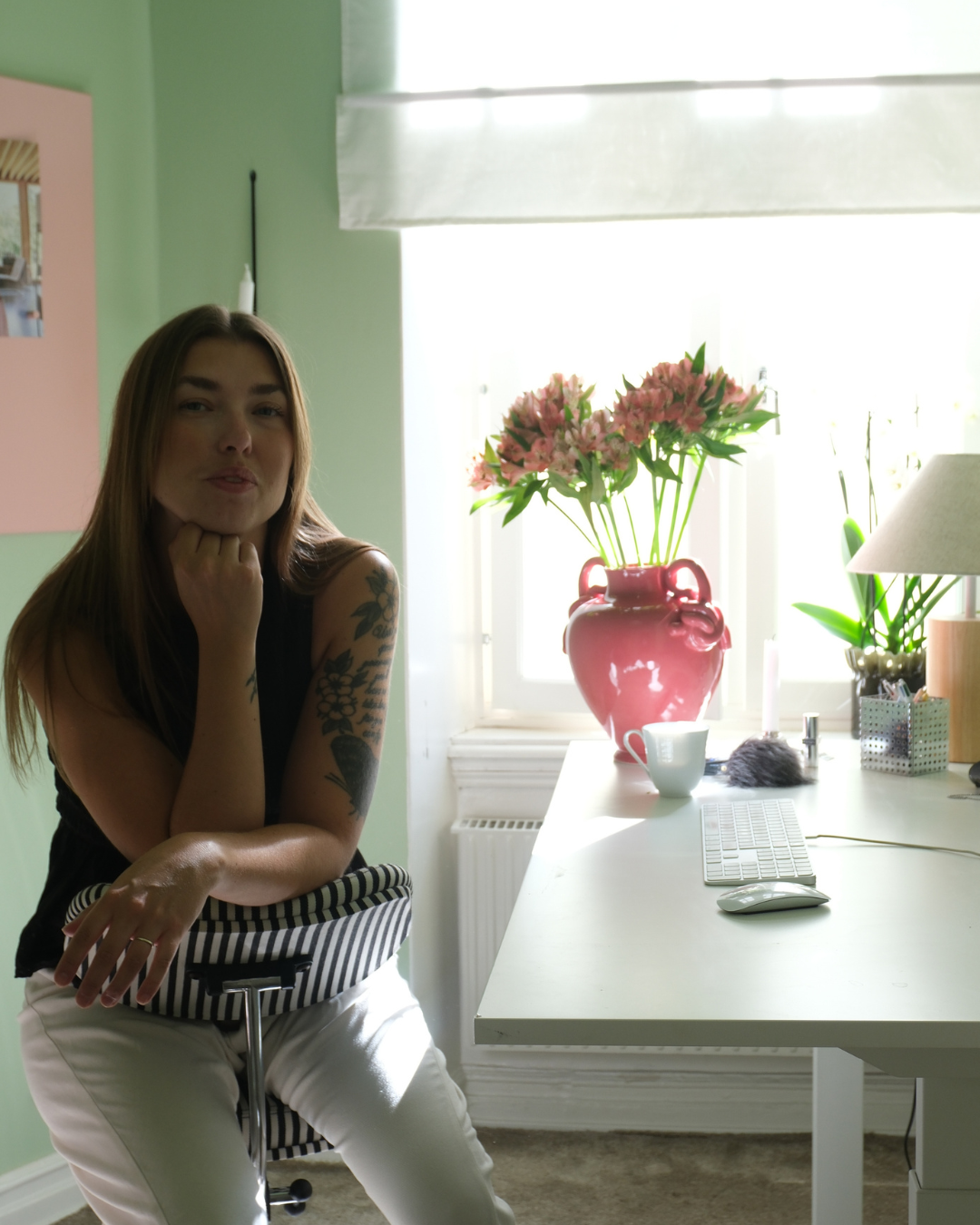 A woman with long brown hair and tattoos on her arm sits at a white desk, resting her chin on her hand, next to a computer keyboard and mouse. Behind her, a window with bright light, a pink vase with pink flowers, and a small white cup are visible on the desk.